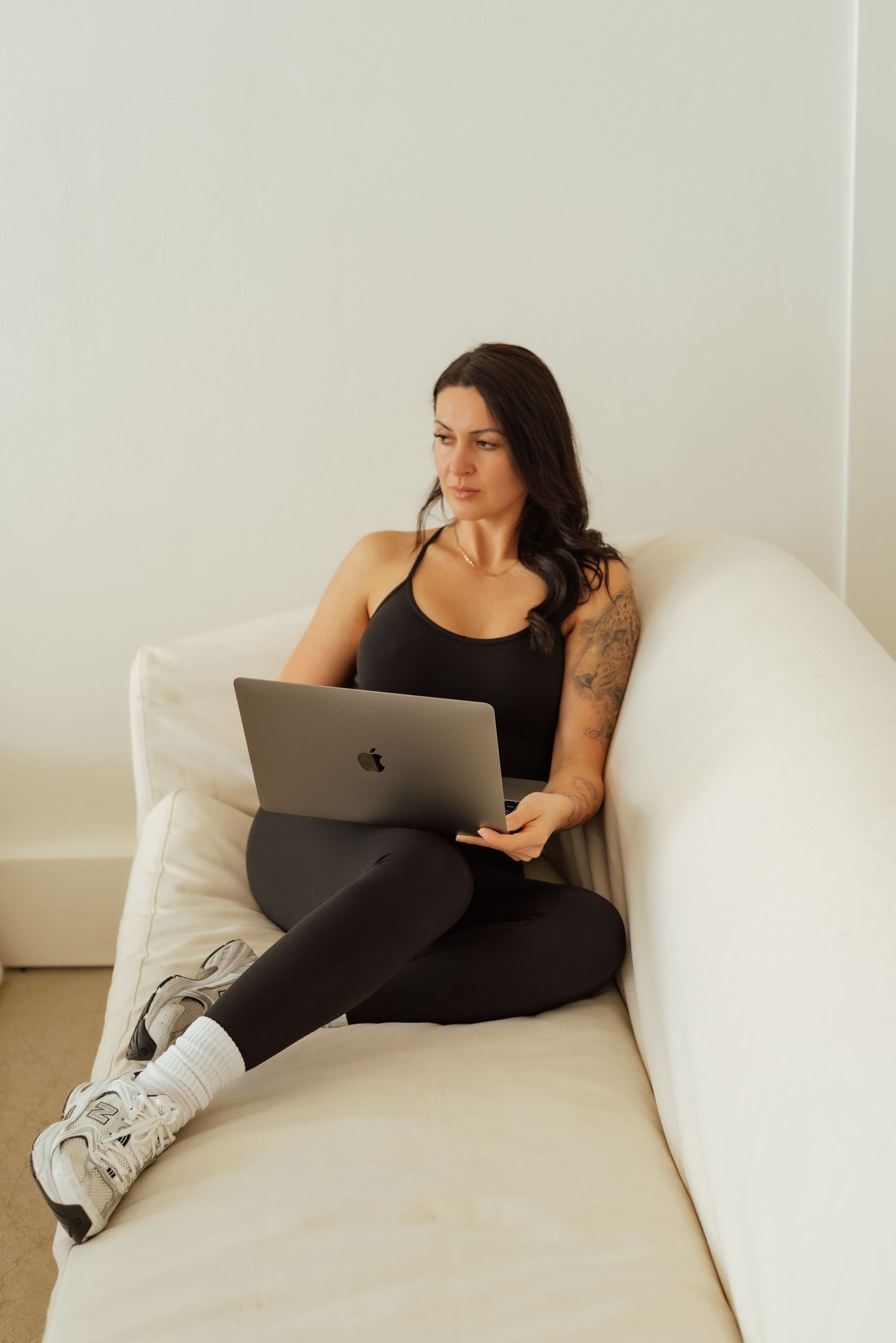 Person in black tank top reclining on white couch while working on MacBook laptop.