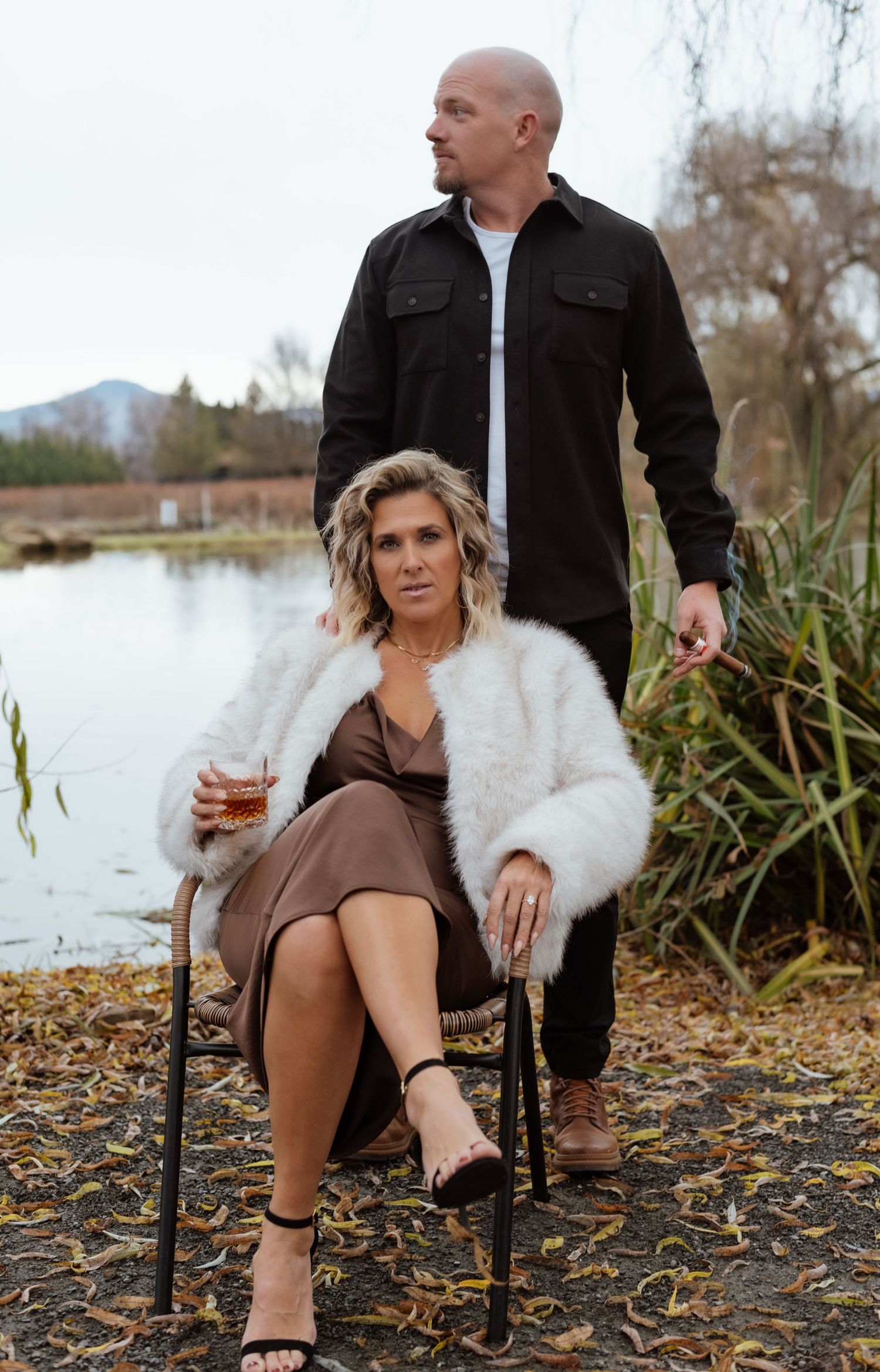 Stylish couple poses by lake, with person in white fur coat seated on vintage chair.