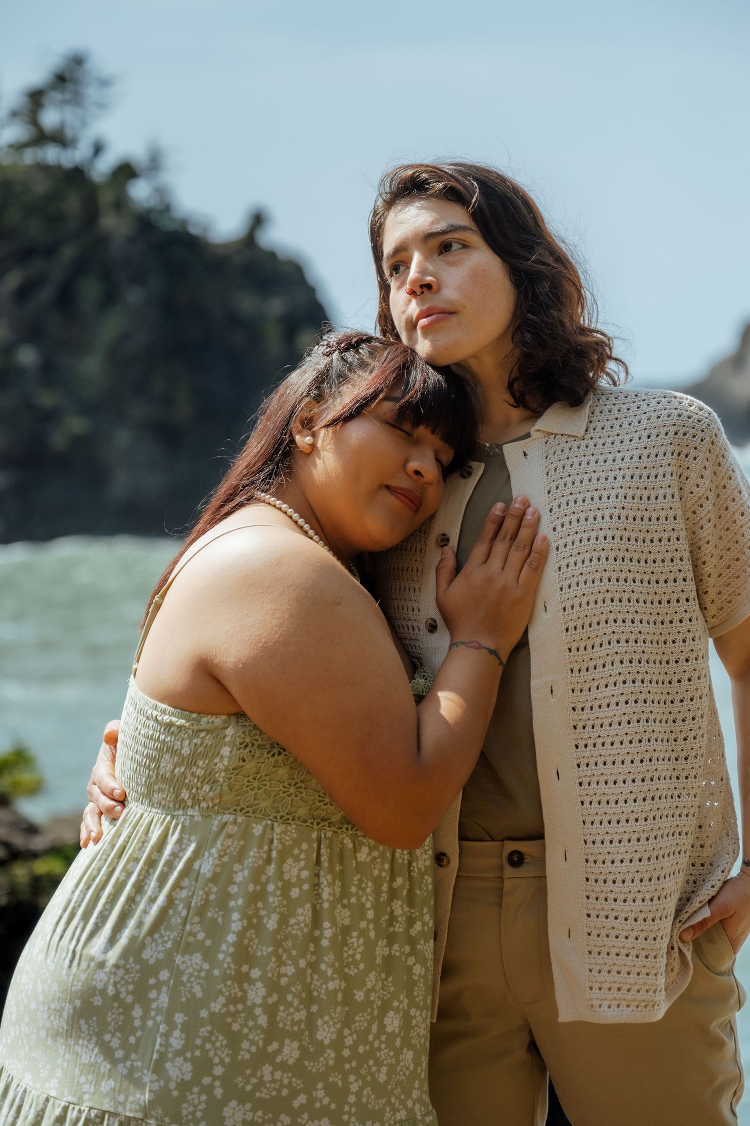 A couple embraces tenderly against a soft blue sky background in matching light-colored outfits.
