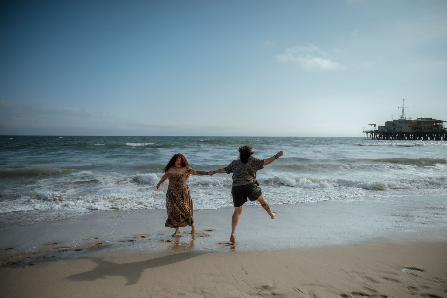 Two people jumping and playing in the ocean waves near a pier on a sunny day.