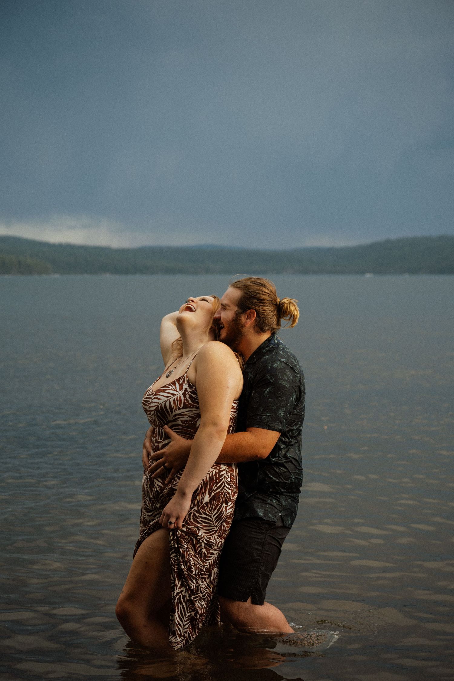 A couple embraces while standing in shallow water with stormy skies and mountains in the background.