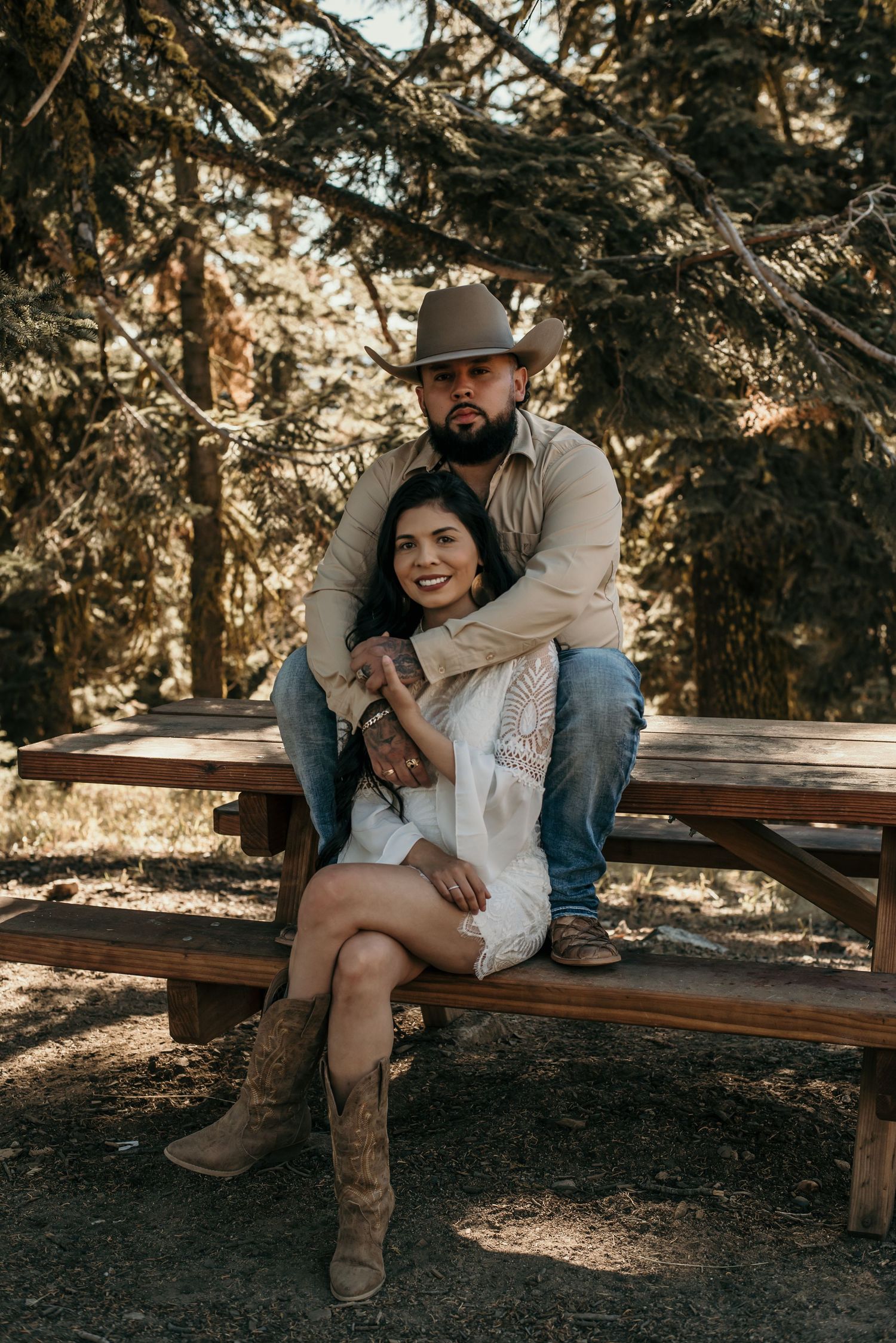 A romantic couple in western attire sits together on a wooden bench in a forest setting.