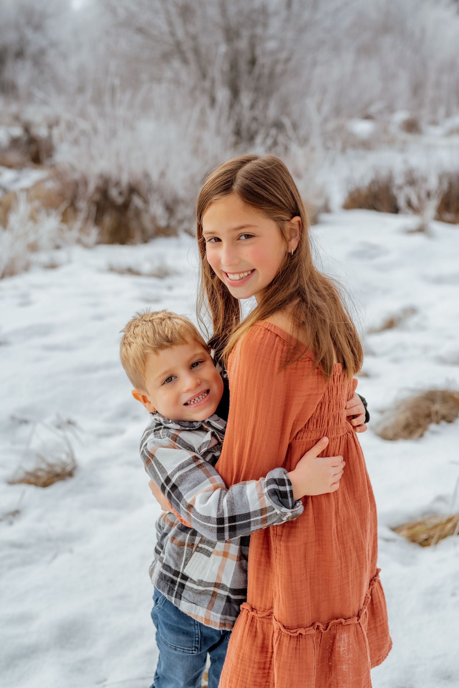 Children share a sweet embrace while wearing orange and plaid in the snowy landscape.
