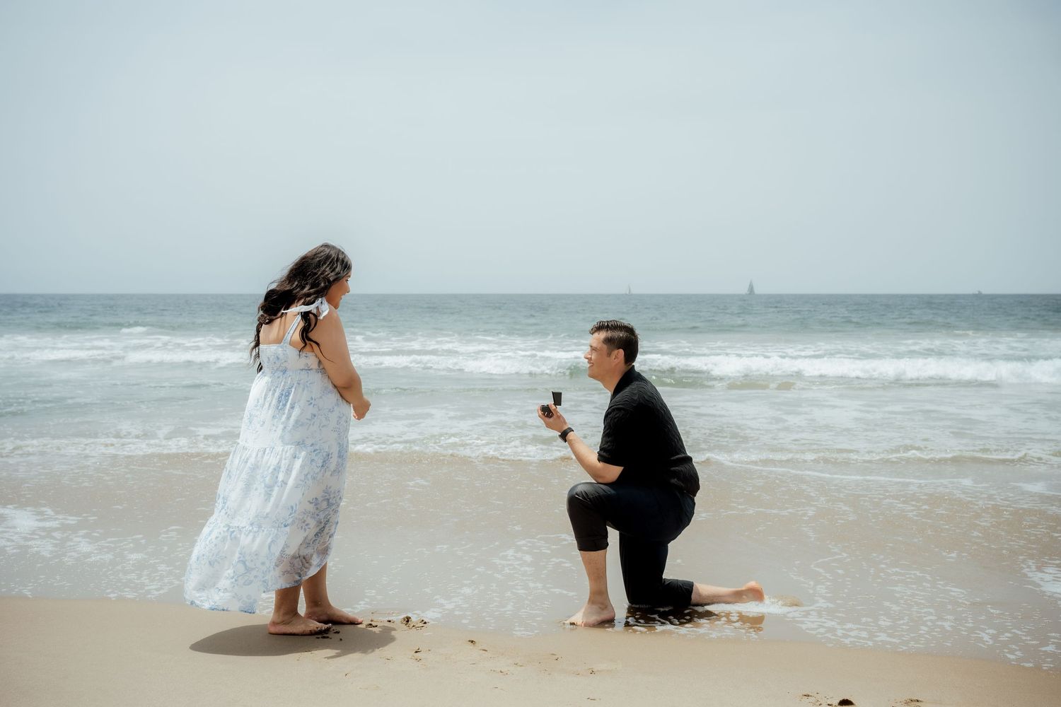 A surprise beach proposal moment captured against ocean waves and cloudy sky.