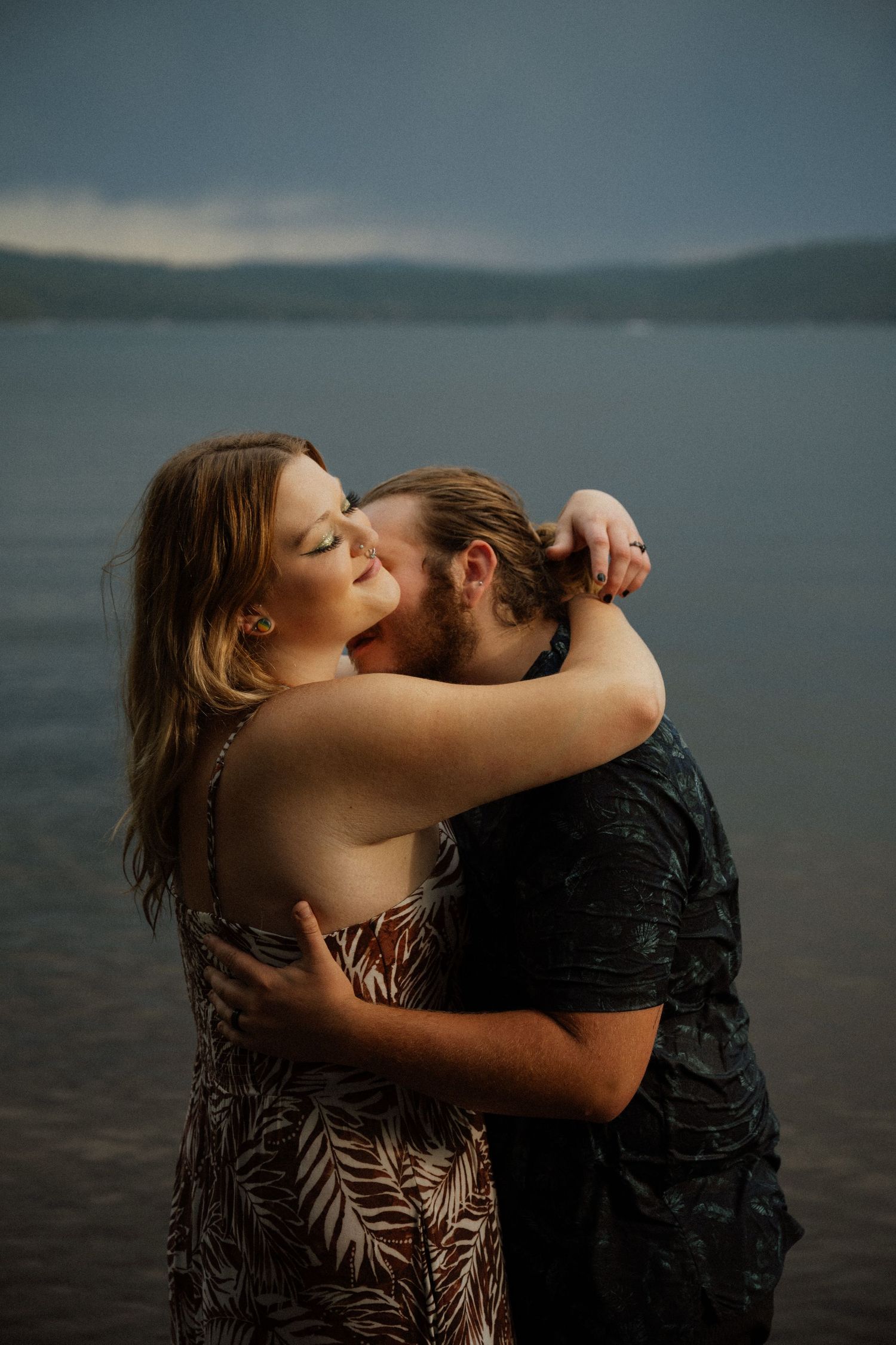 Couple sharing an intimate moment by a serene lake with mountains visible in the distance.
