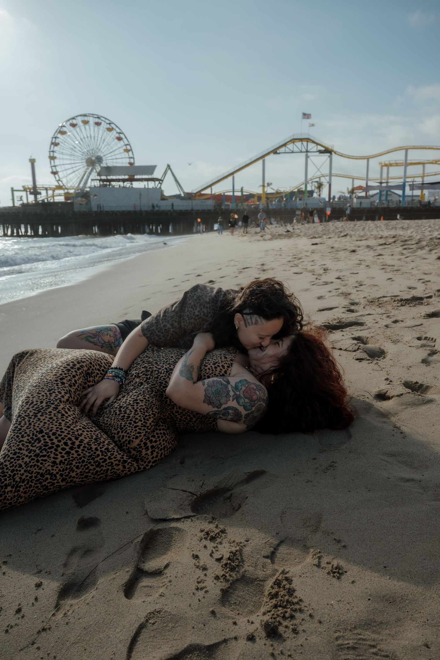 Person lying on dark sand beach with amusement park pier and rides in background.