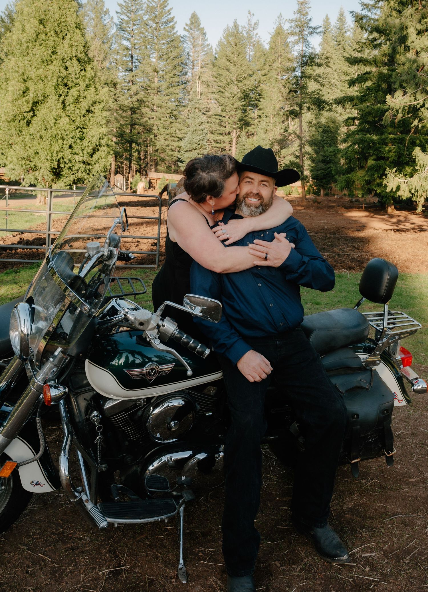 A couple sharing an intimate moment on a motorcycle in a wooded setting.