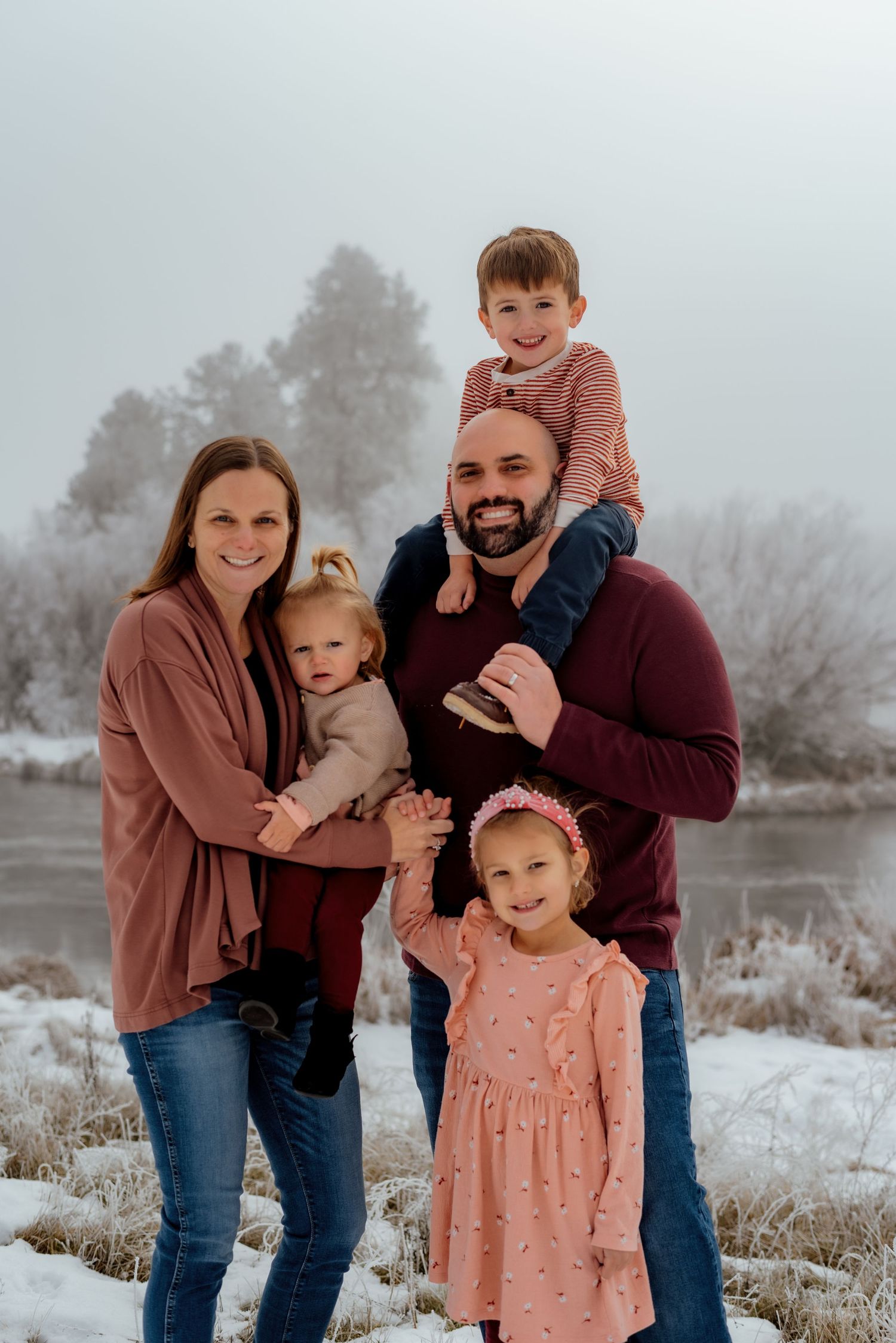 Family of four posing together in coordinated outfits against snowy winter landscape.