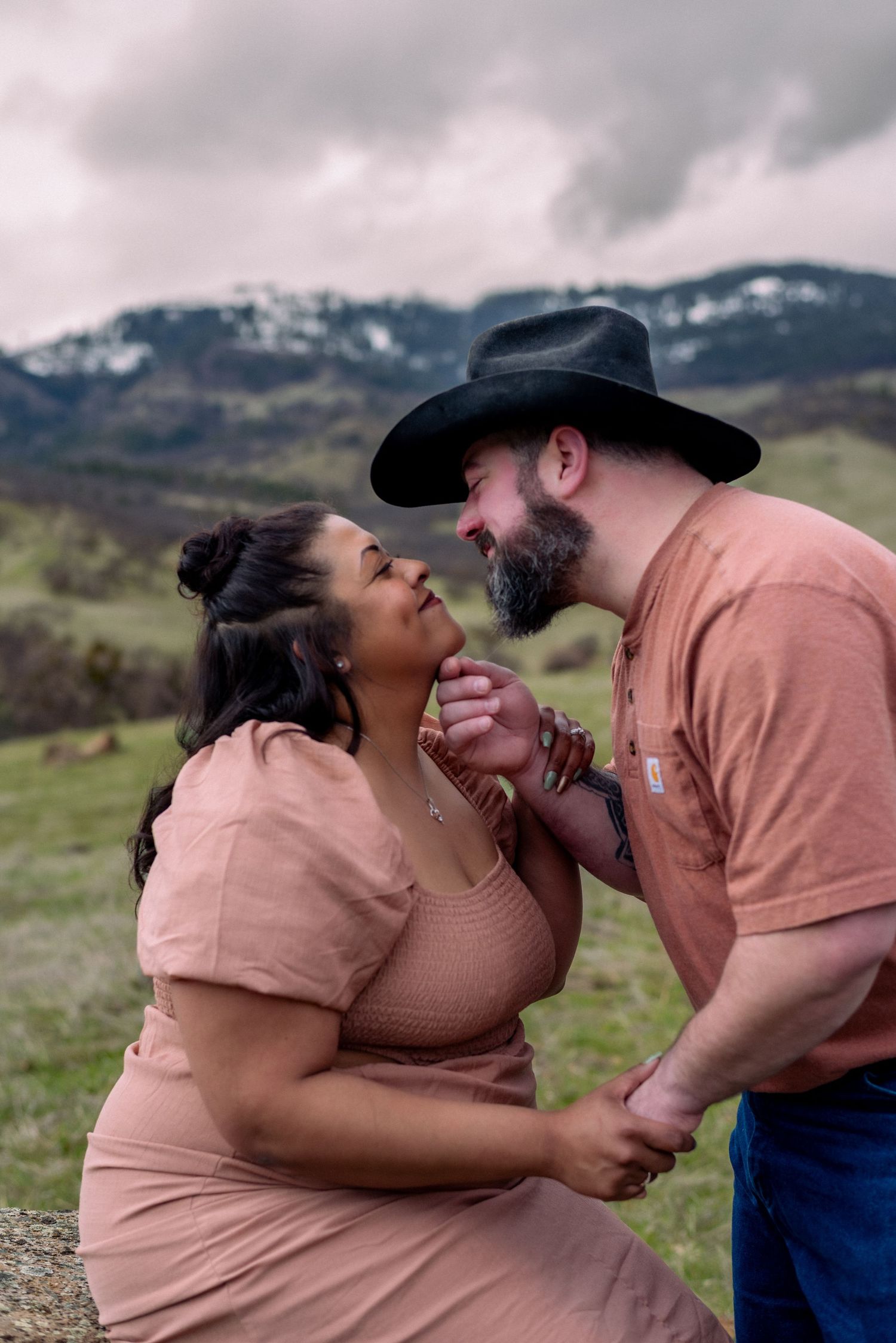 A romantic couple shares tender moments against a cloudy mountain backdrop.