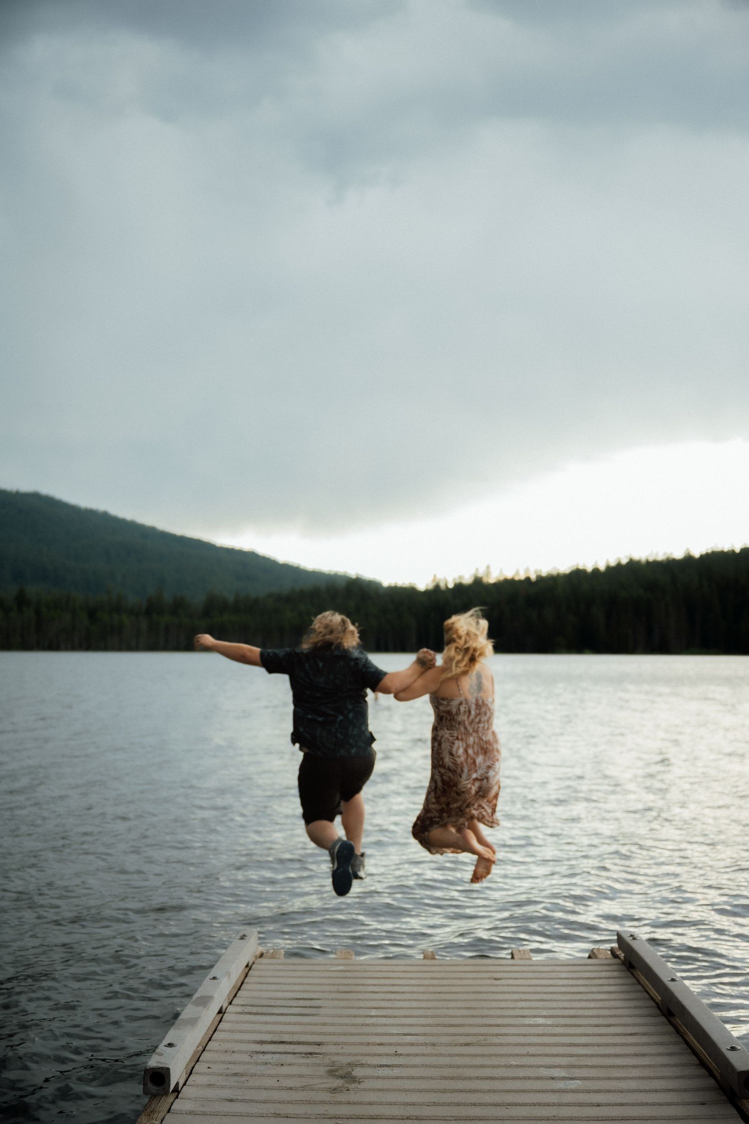 Two people jumping off a wooden dock into a mountain lake with dramatic cloudy skies.