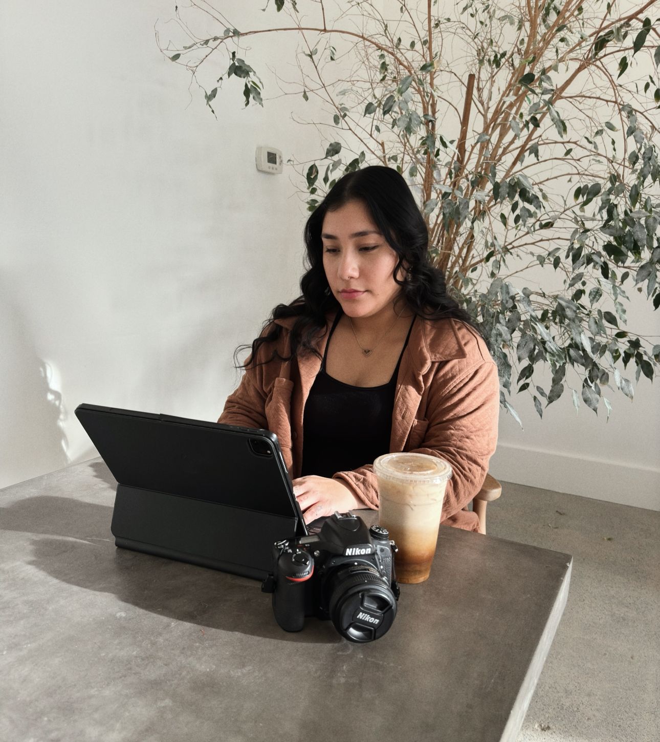 A person works on a tablet with a camera and coffee in a minimalist white room with dried flowers.