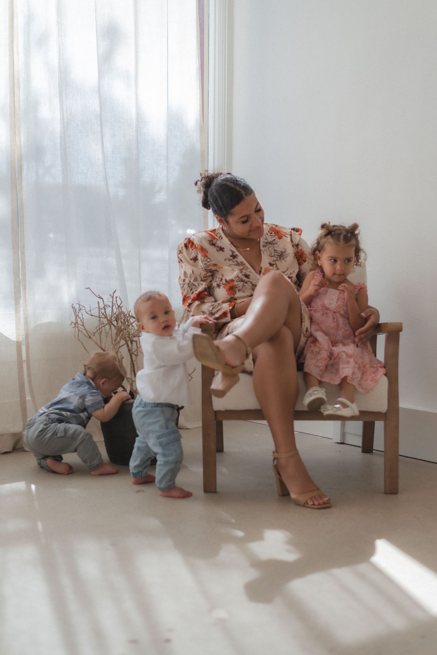 Family photo session with children and adult posing playfully near wooden chair by window.