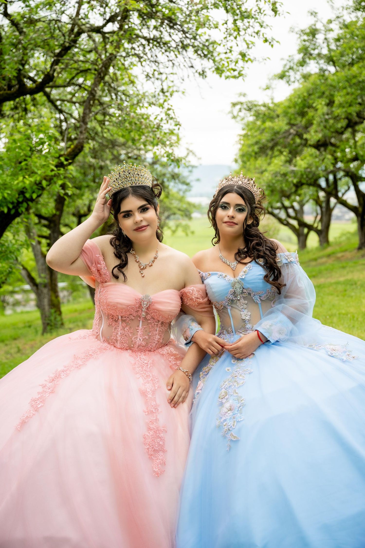 Two elegant ballgowns, one in pink and one in blue, photographed in a scenic orchard setting.
