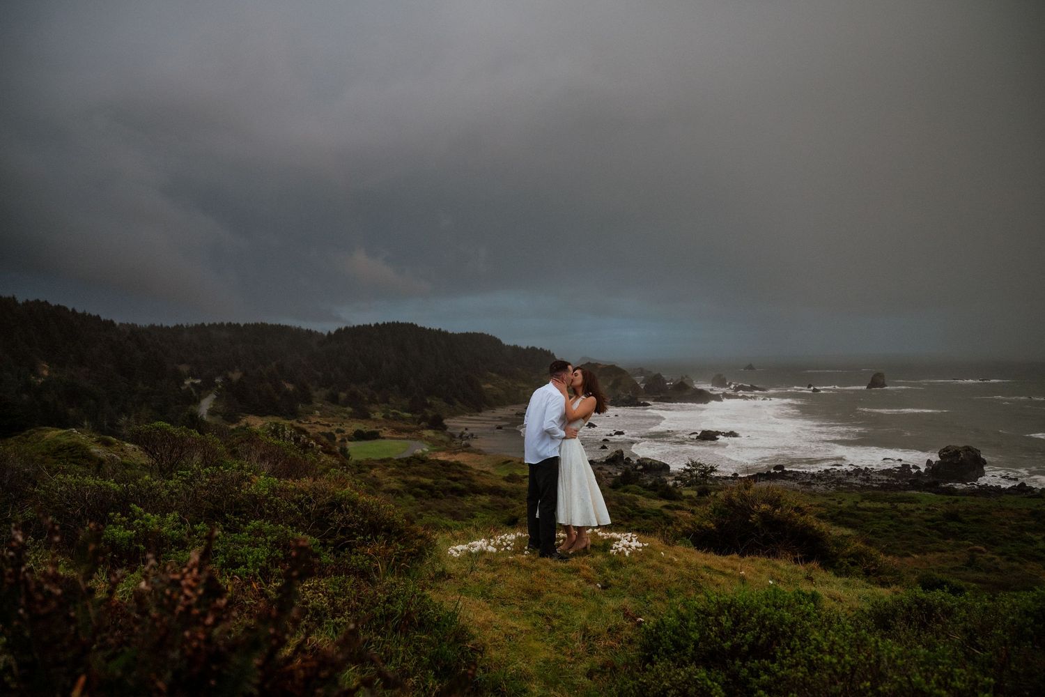 A couple embraces on a dramatic coastal cliff under stormy skies.