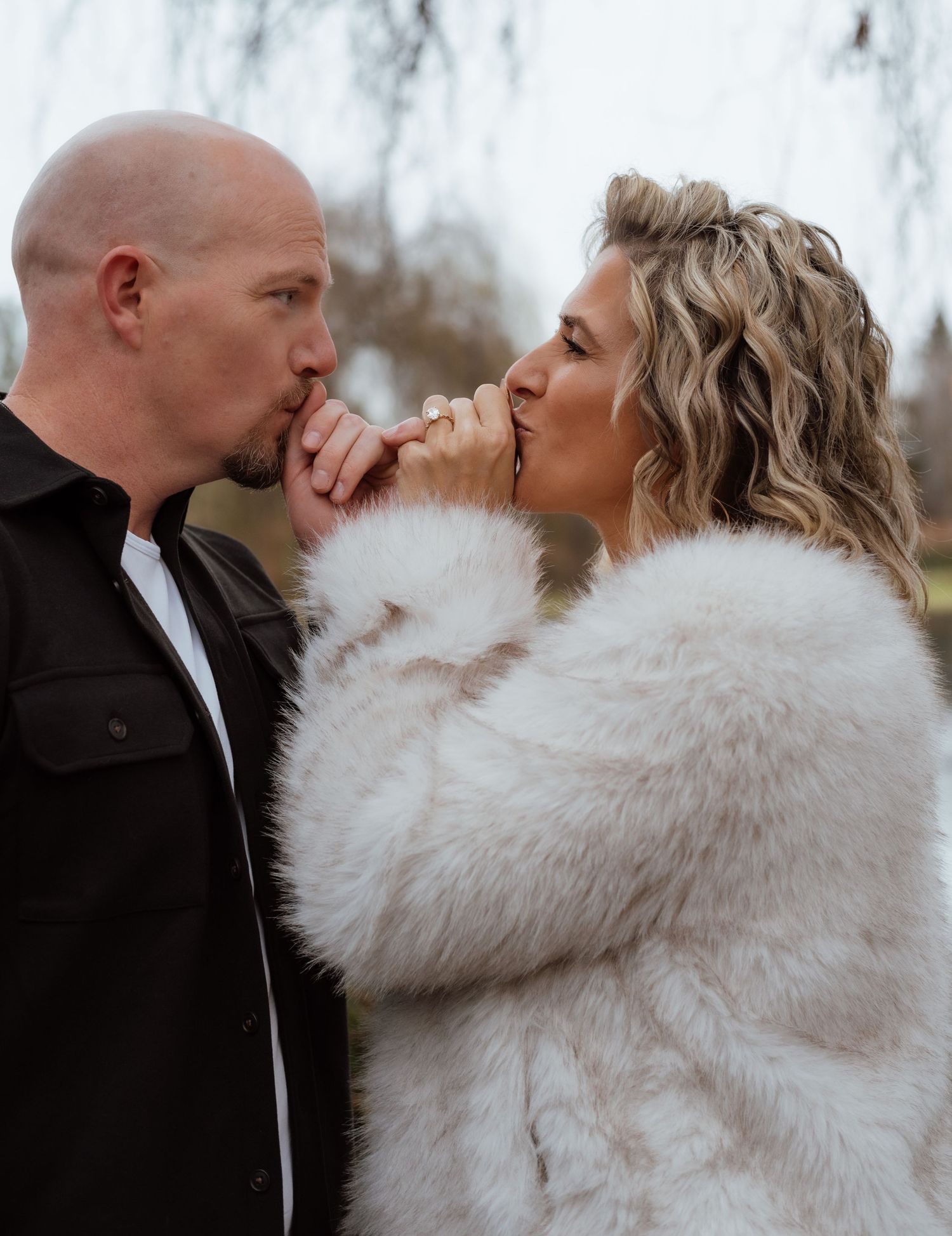 Intimate moment between couple with person wearing white fur coat against willow tree background.