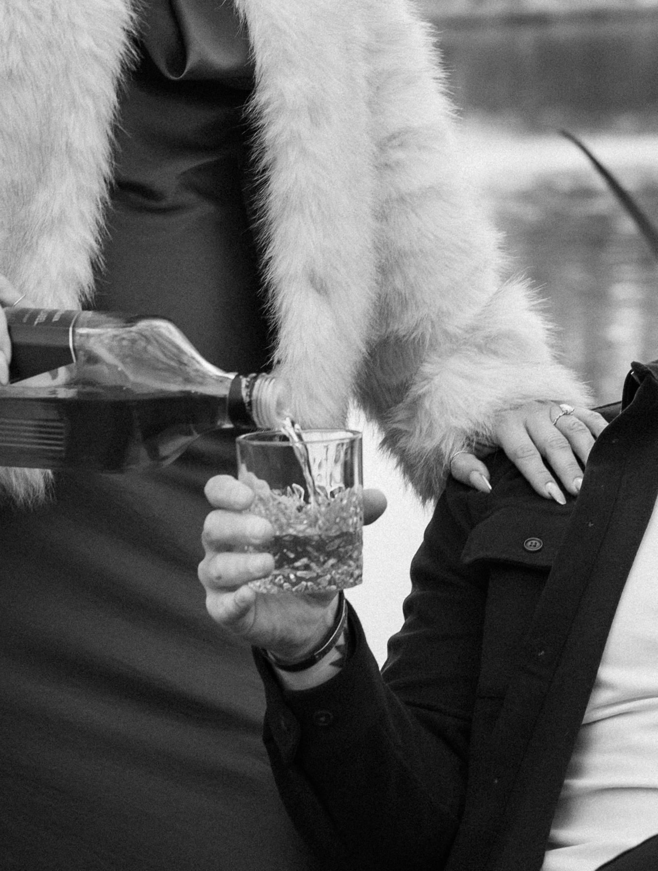 Close-up of hands pouring drinks in crystal glasses, with a white fur coat visible.