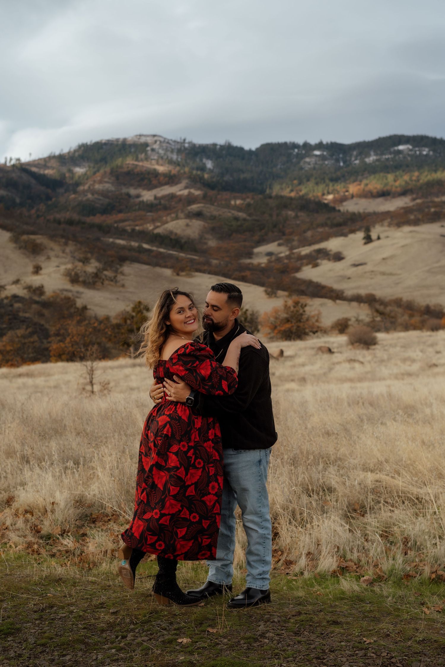 Couple embracing in a red dress and casual wear against rolling autumn hills.