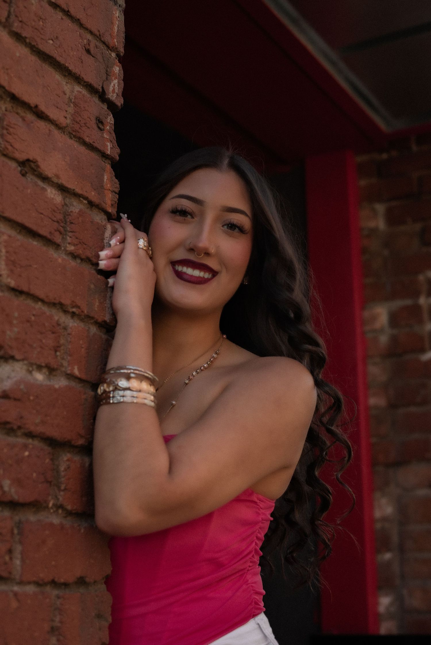 Glamorous portrait with pink top and statement jewelry against red brick background.
