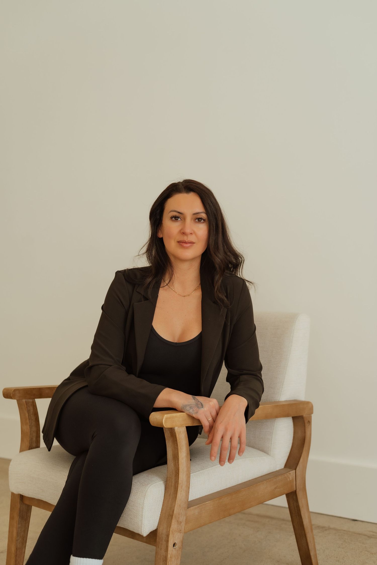 Professional woman in black business attire seated in wooden chair against white backdrop.
