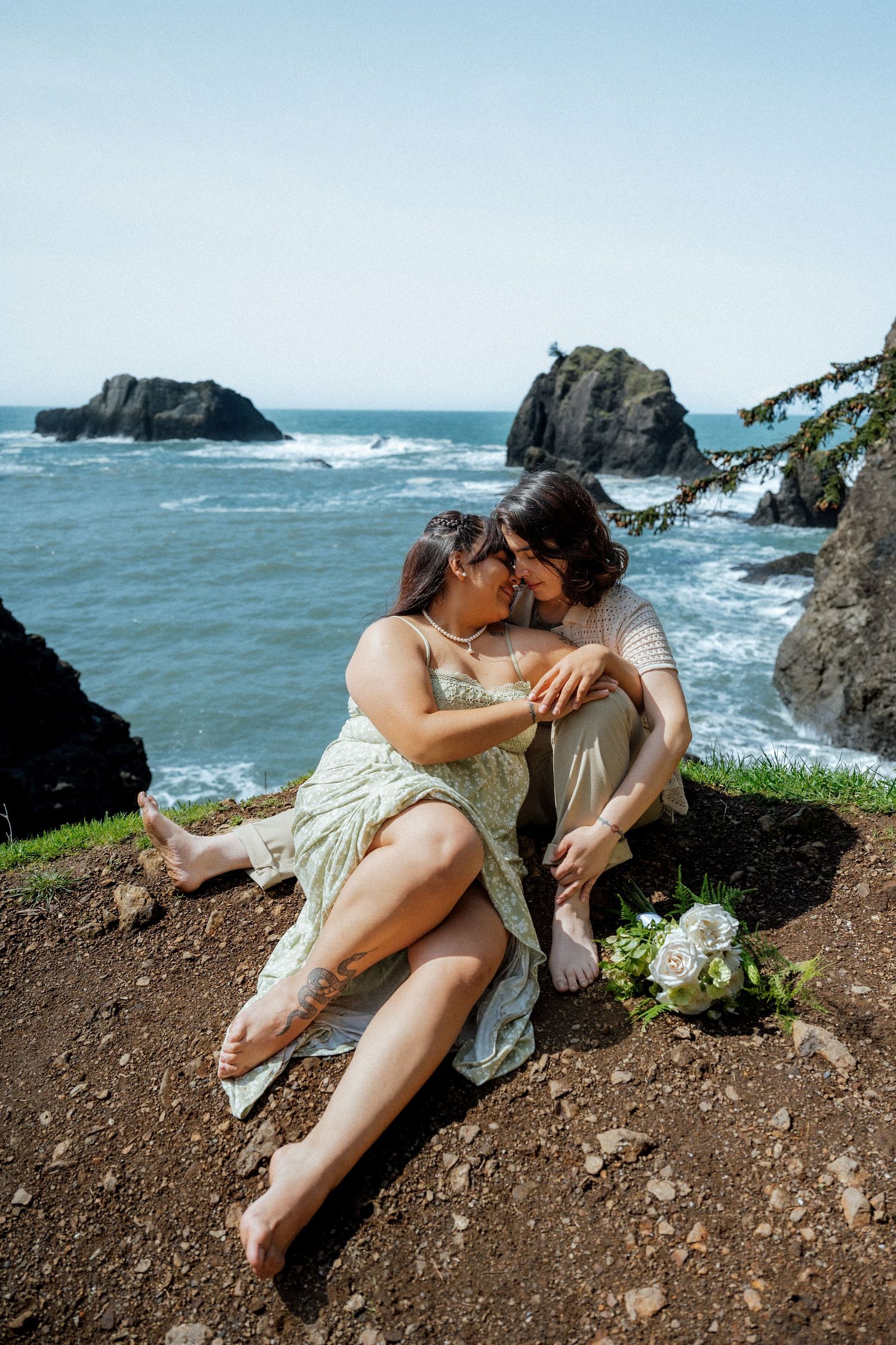 Two people share an intimate moment on a cliff edge with dramatic ocean views and rock formations.