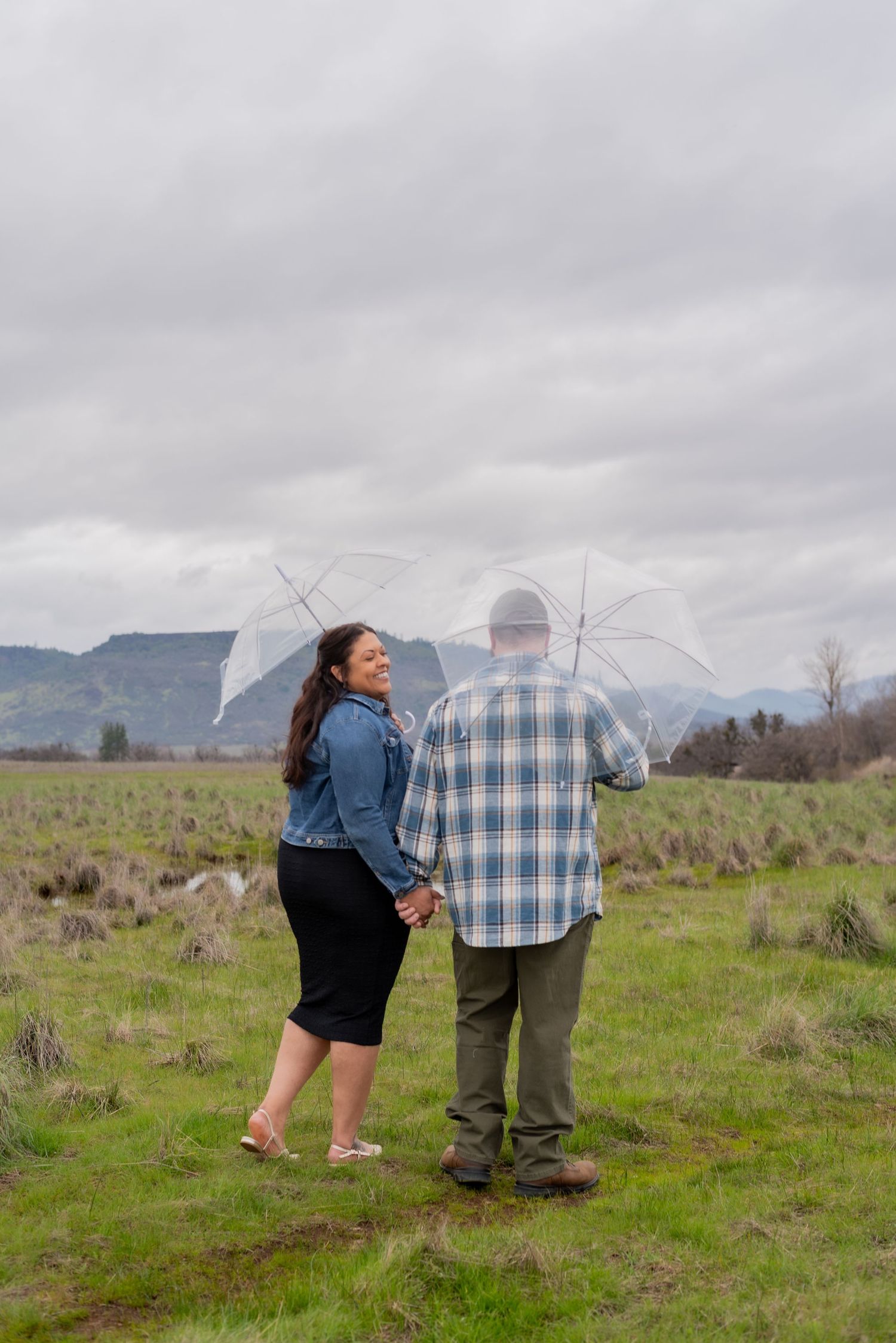 A couple stands together in a scenic field with mountains in the background.