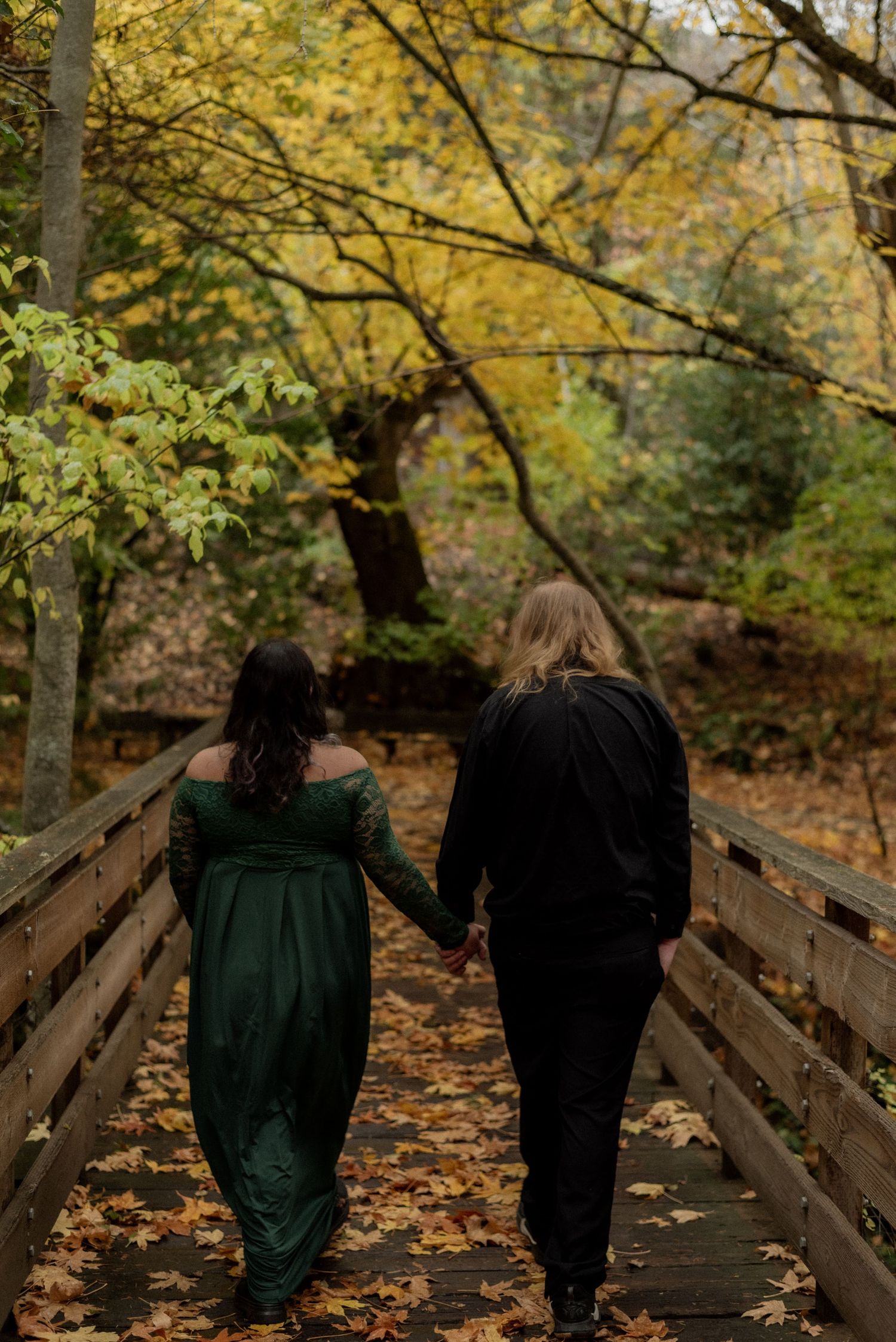 A couple walks hand in hand on a wooden bridge surrounded by fall foliage.