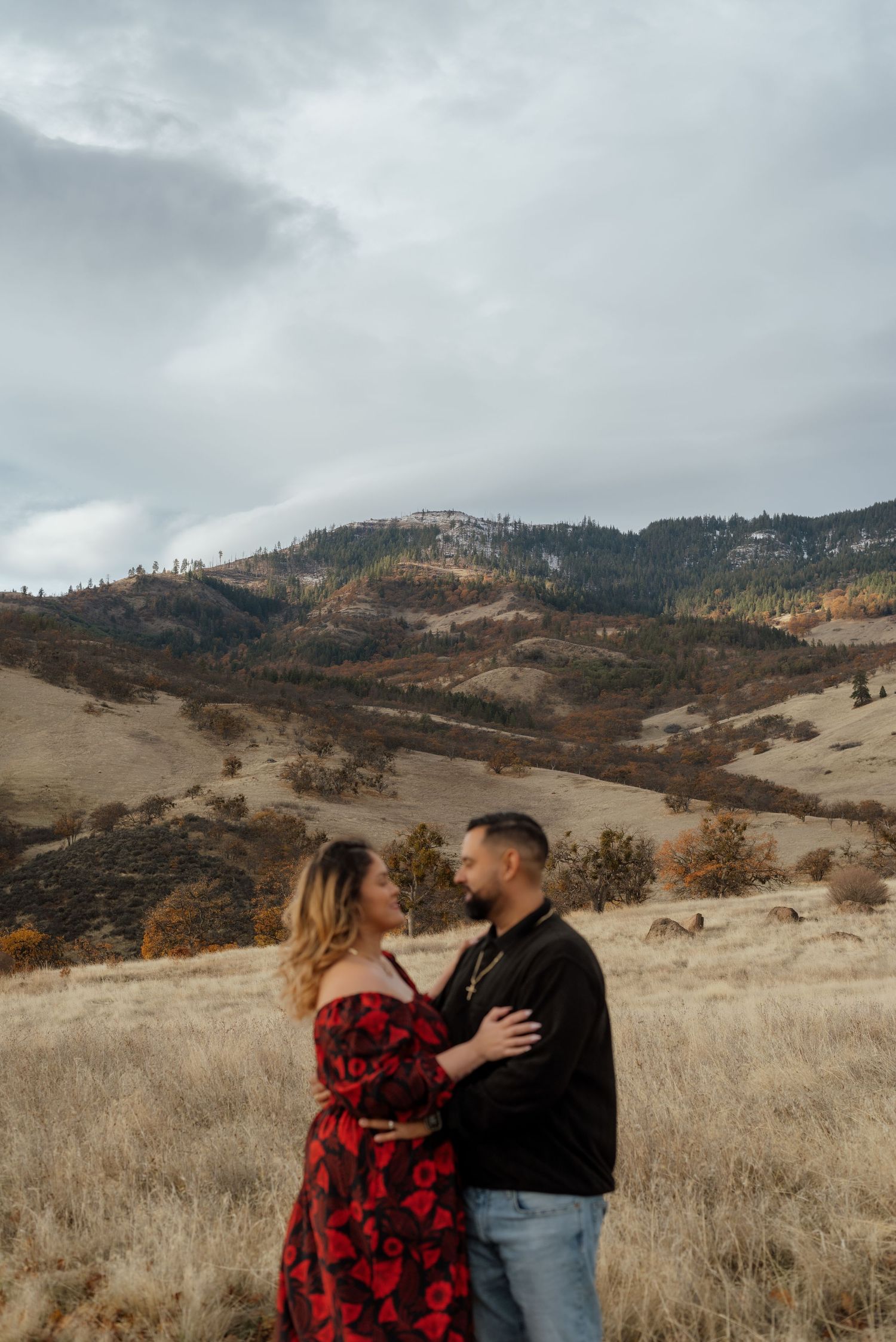 Couple in casual wear embracing in a dry grass field with rolling hills behind them.