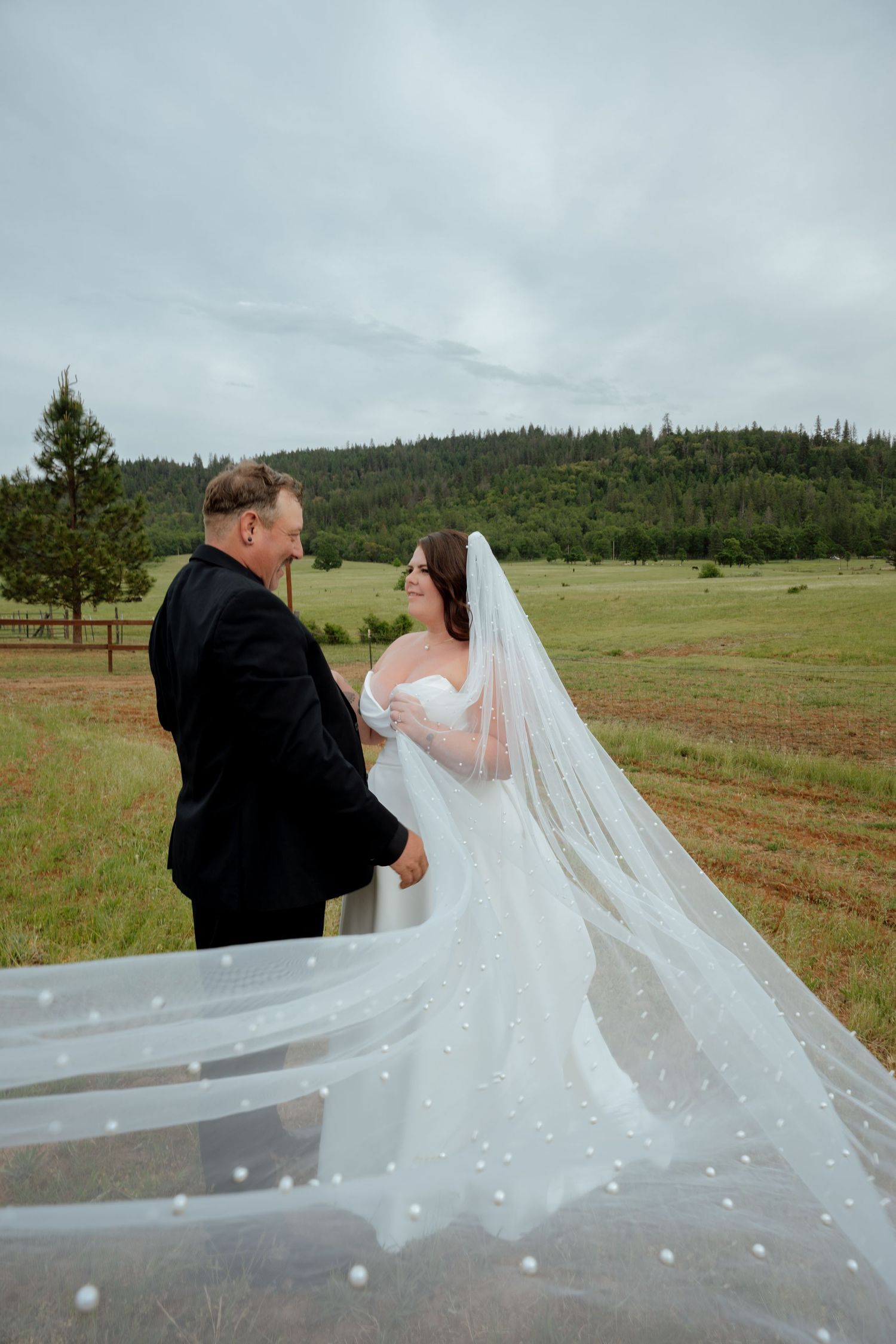 A bride's flowing veil catches the wind as couple shares a moment in a mountain meadow.