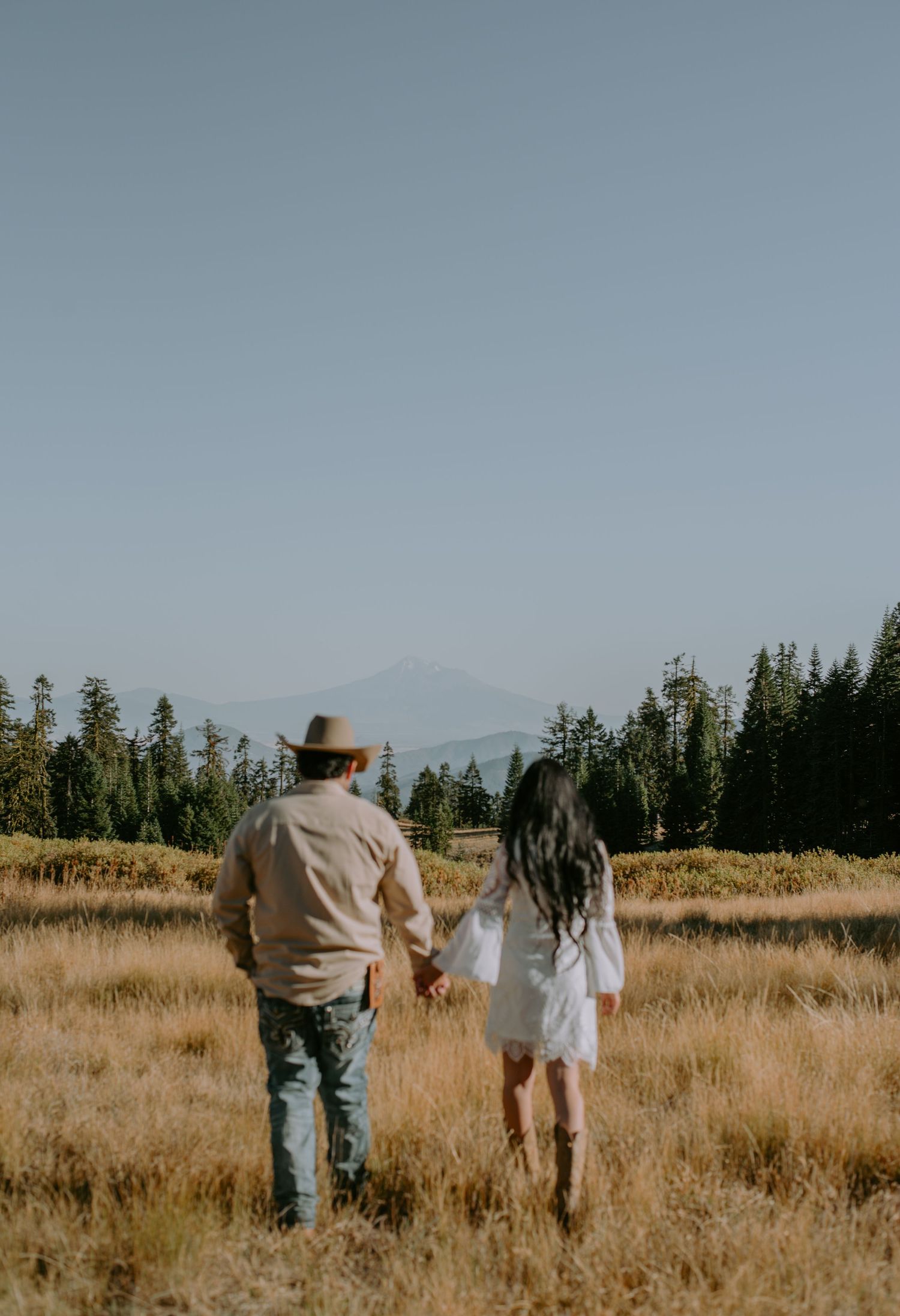 A couple walks hand in hand through a golden field with mountains in the background.