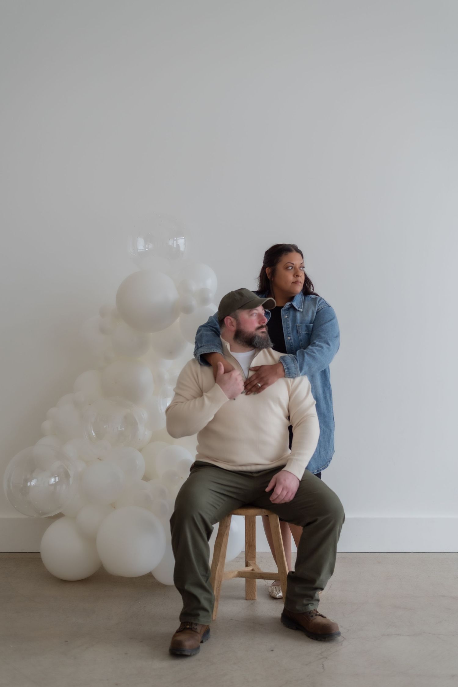 A person sits alone on a wooden stool against a white wall with white balloons in the background.