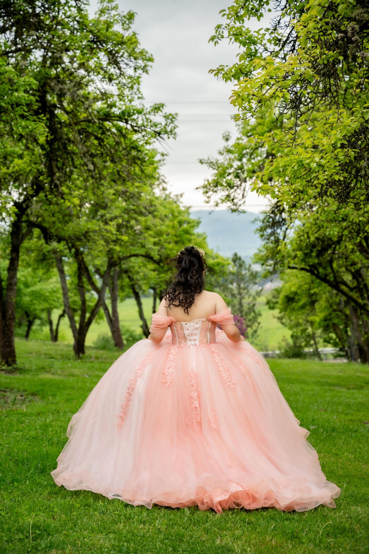 Stunning peach princess ball gown photographed against green trees in a garden setting.