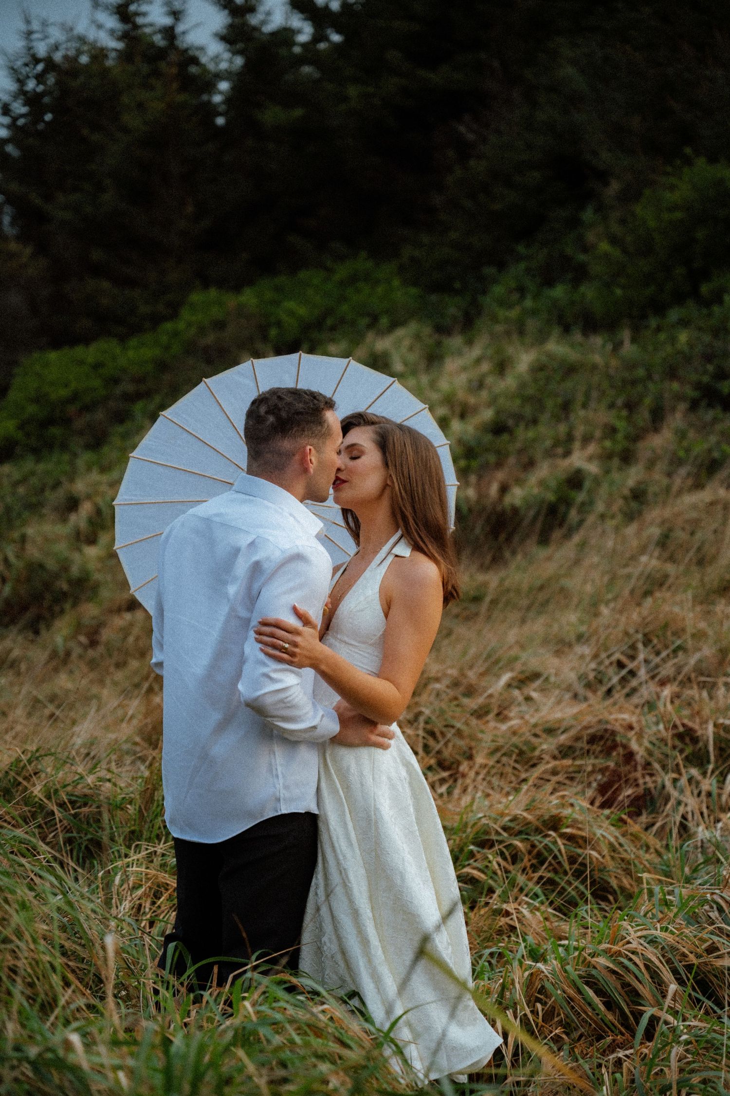 A couple kisses while holding a white umbrella on a grassy hillside.