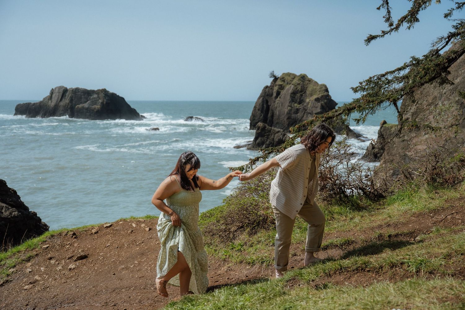 A couple holds hands while hiking along a scenic coastal cliff with rocky ocean formations in the background.