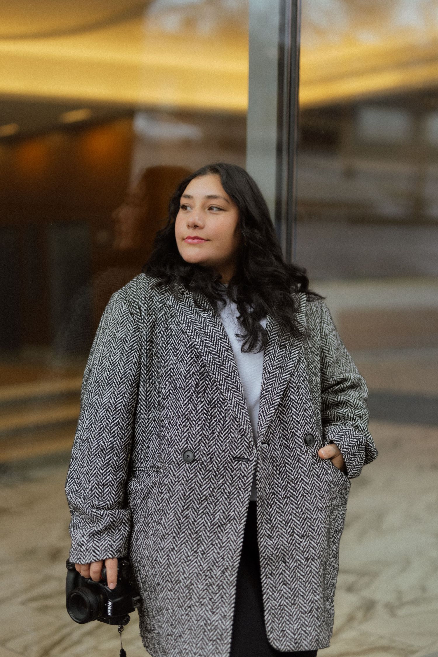A person in a grey wool coat poses confidently against a modern glass building.