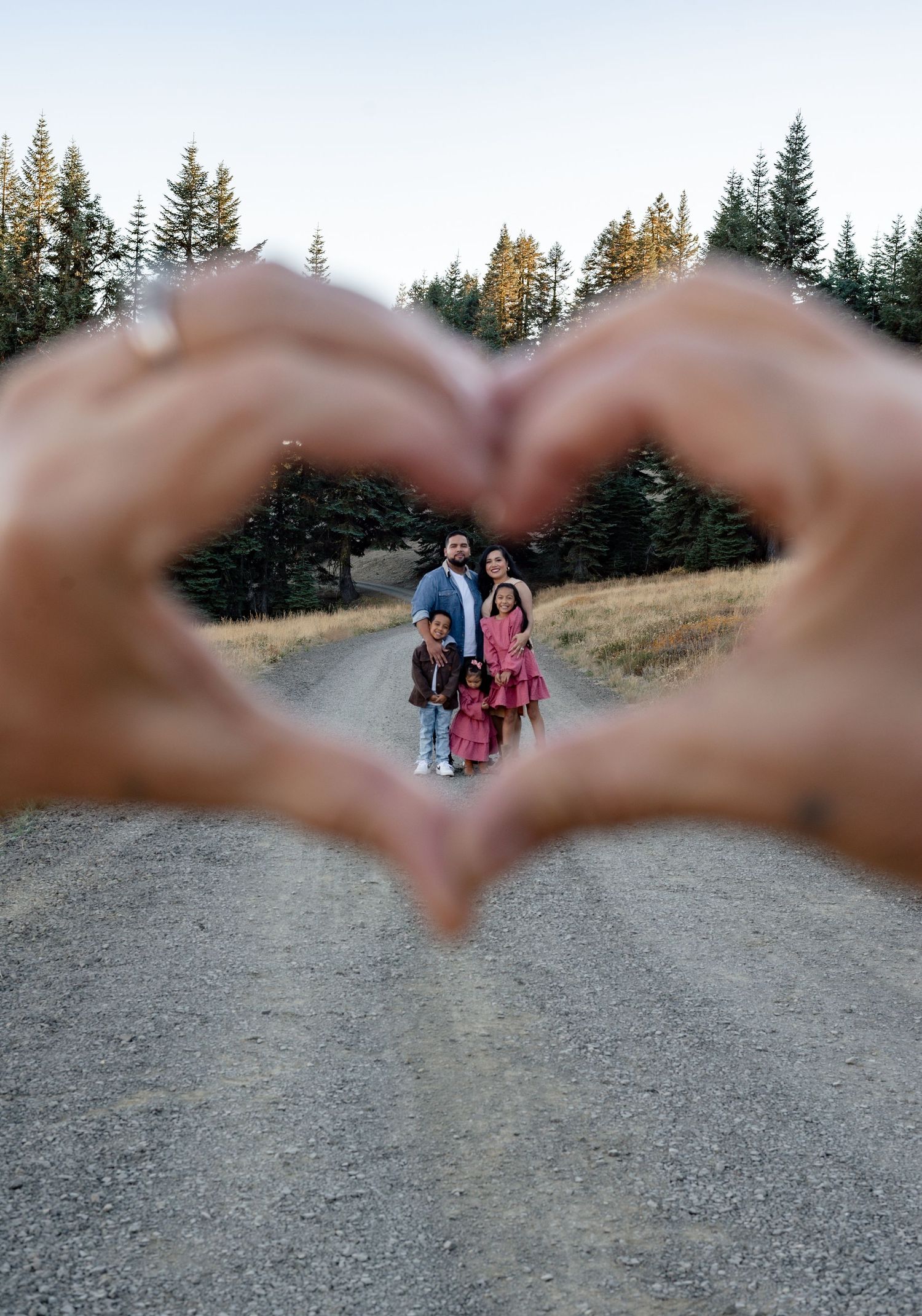 Hands forming a heart shape framing a family on a mountain path at sunset.