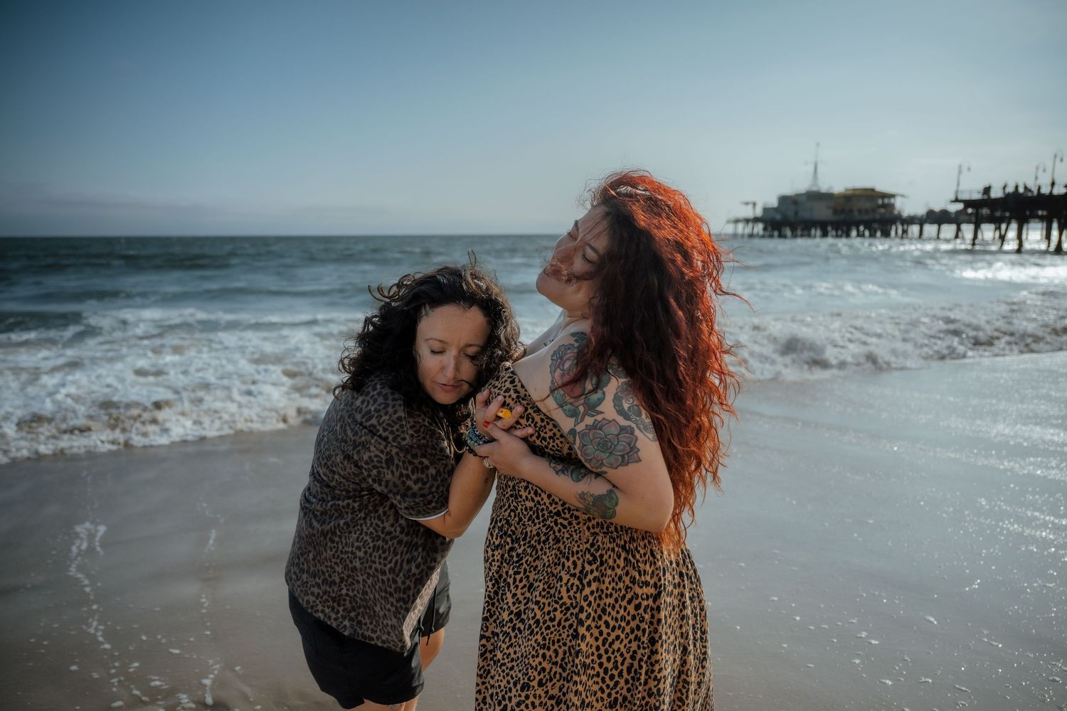 Two people in animal print clothing laughing together on a beach with a pier in the background.