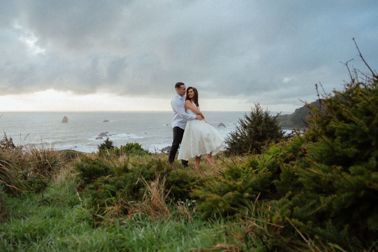 A couple shares an intimate moment on a windswept coastal bluff with dramatic clouds.