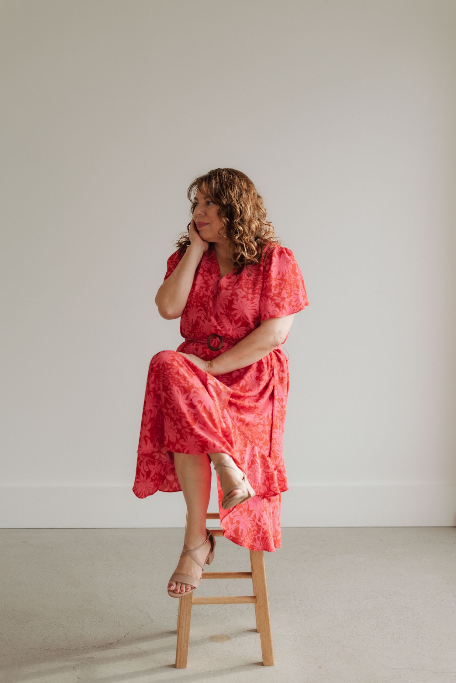 Woman in flowing coral dress poses playfully on wooden stool against white backdrop.