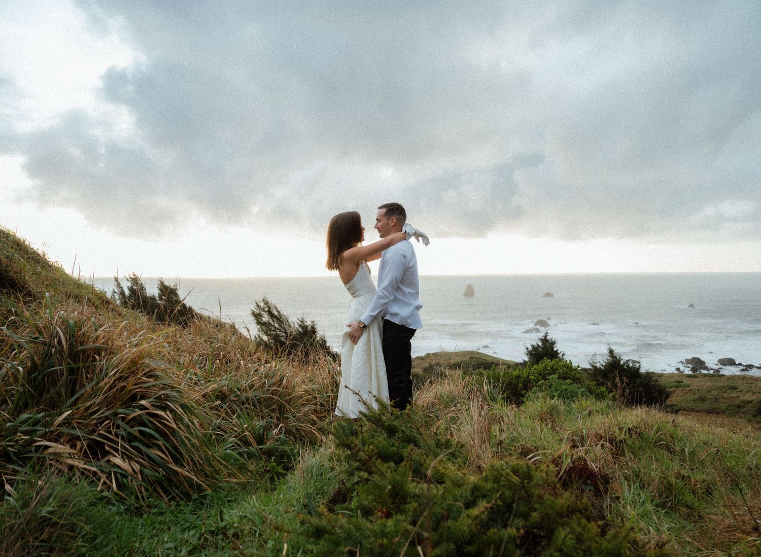 A couple embraces on a coastal clifftop overlooking the ocean under moody skies.
