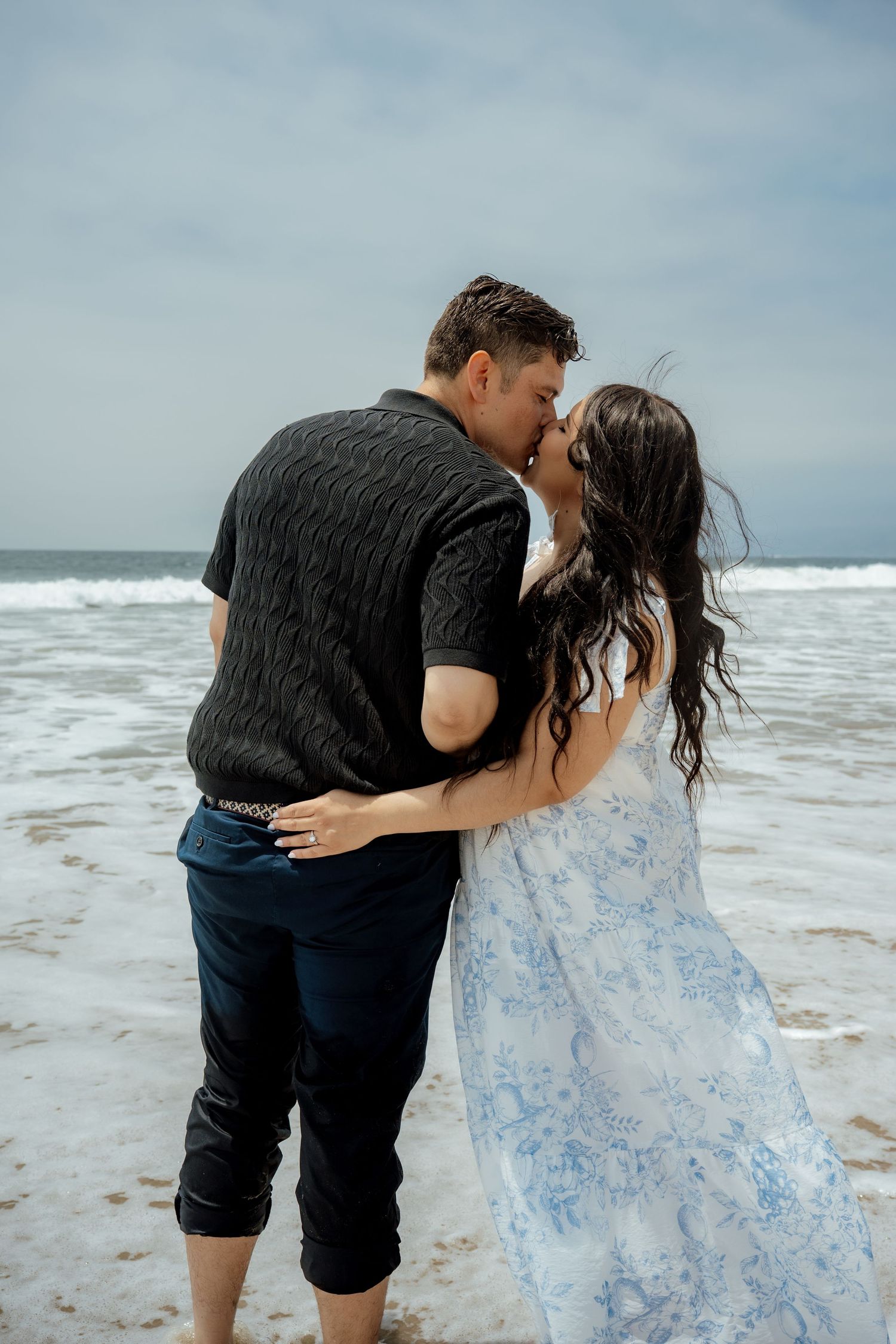 A romantic couple embraces while standing on a sandy beach with crashing waves.
