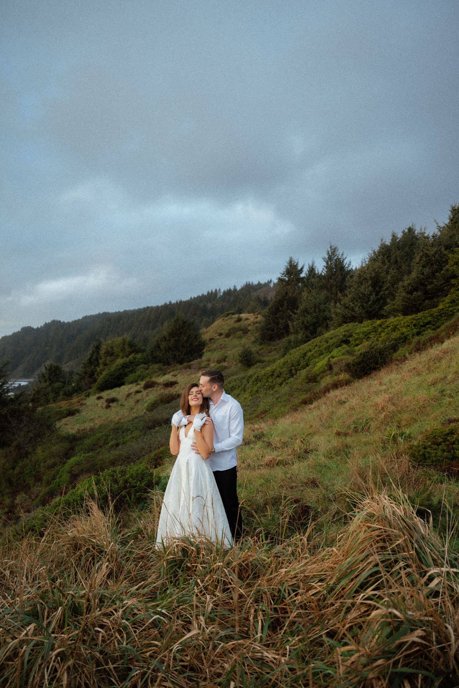 A romantic moment captured on a coastal hillside with tall grass and cloudy skies.