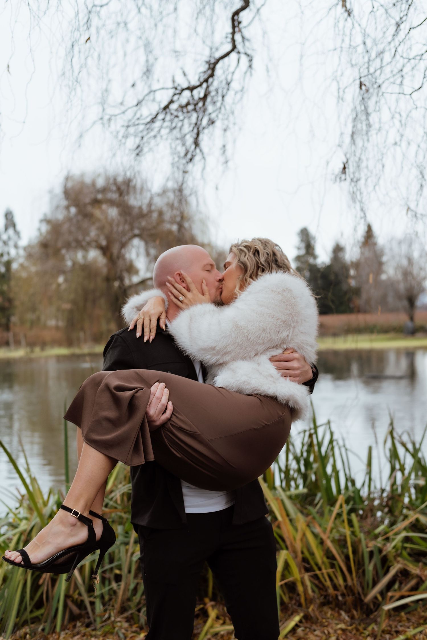 Romantic couple embracing by lakeside with person in brown dress being lifted.