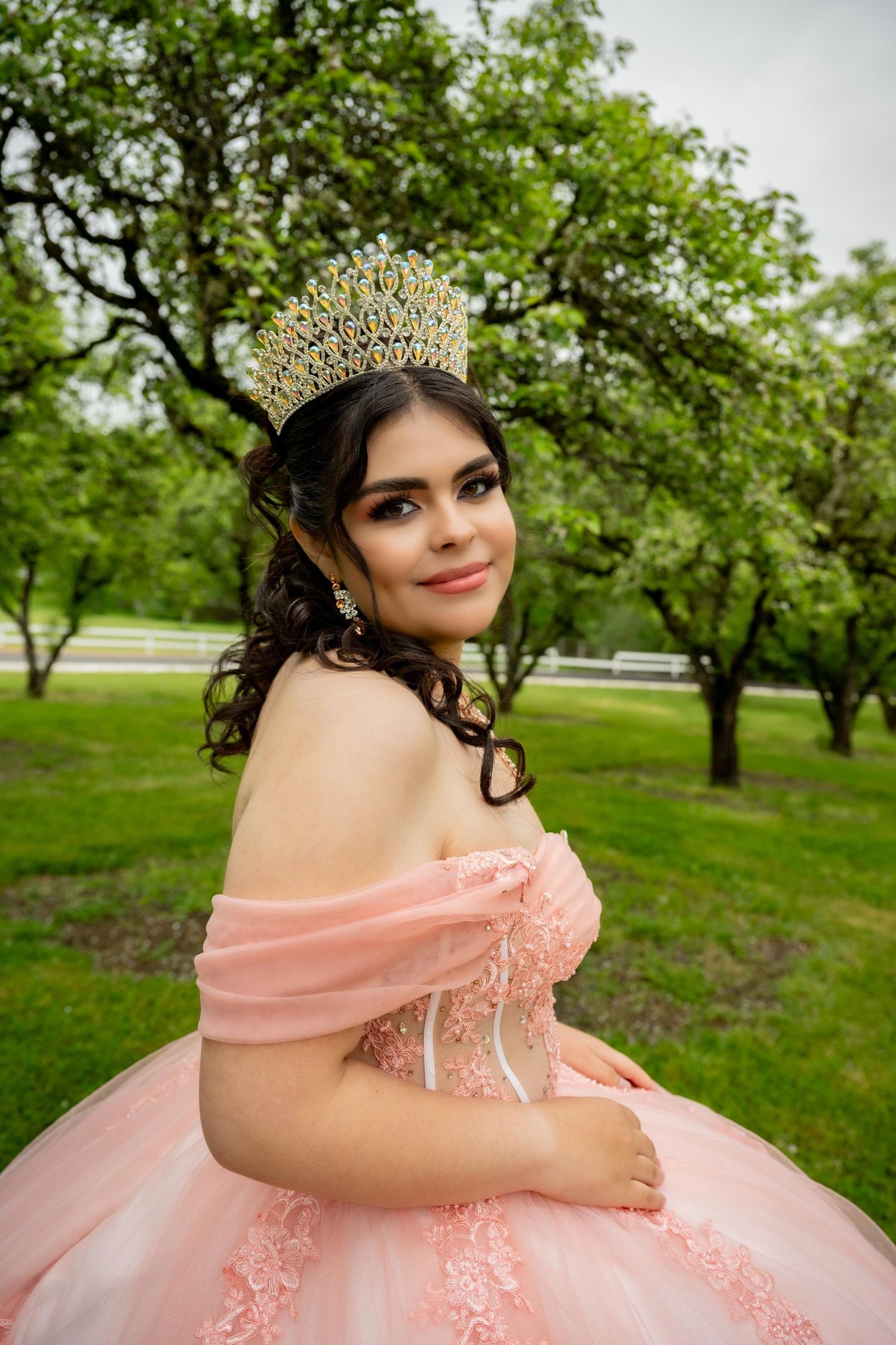 Elegant peach quinceañera gown with off-shoulder design photographed in a lush green garden setting.