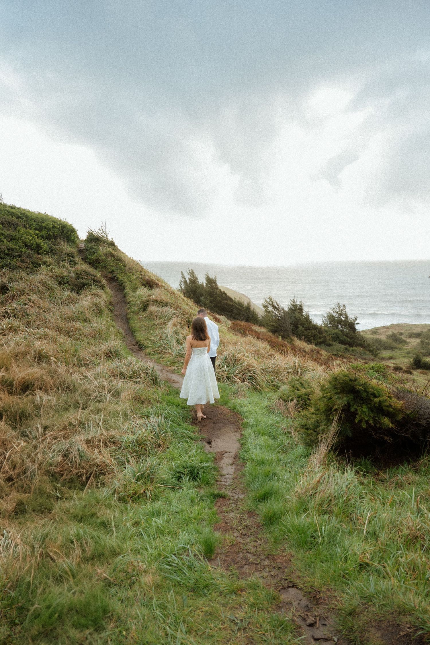 A person in a white dress walks along a coastal hiking trail with ocean views.