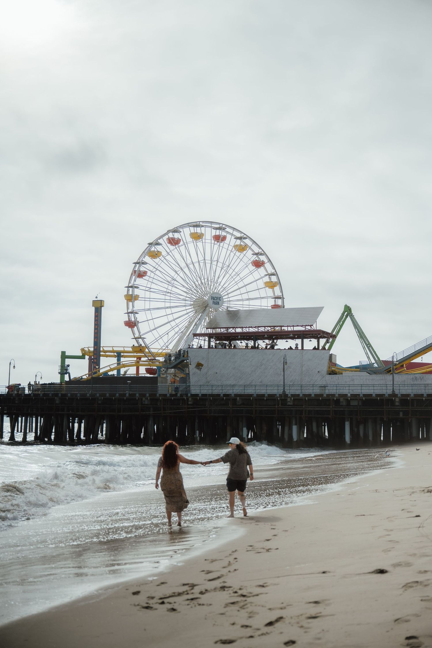 Two people walk hand in hand on a beach with a Ferris wheel on a pier in the background.