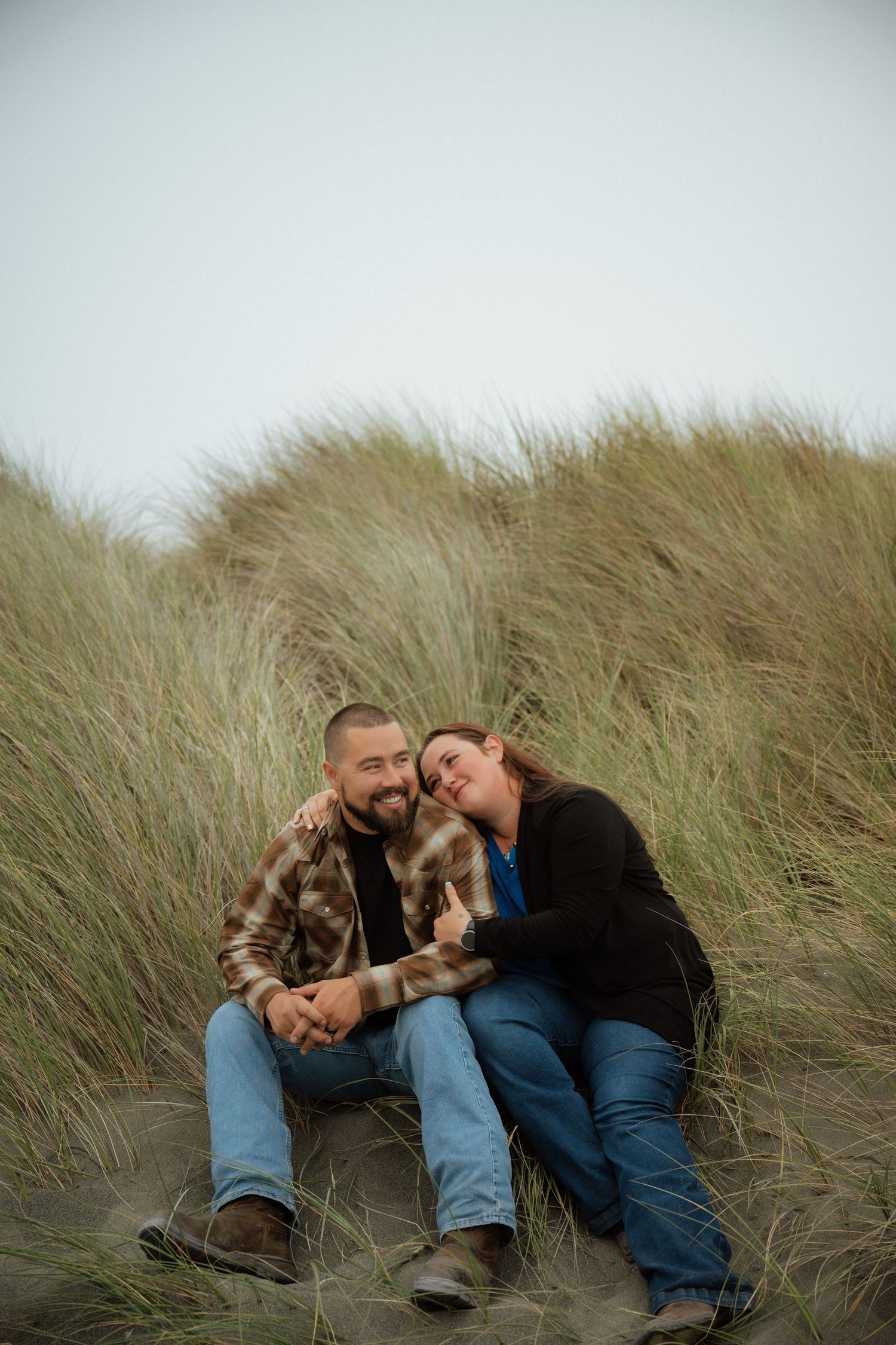 Two people rest together among coastal dune grass on a cloudy day.