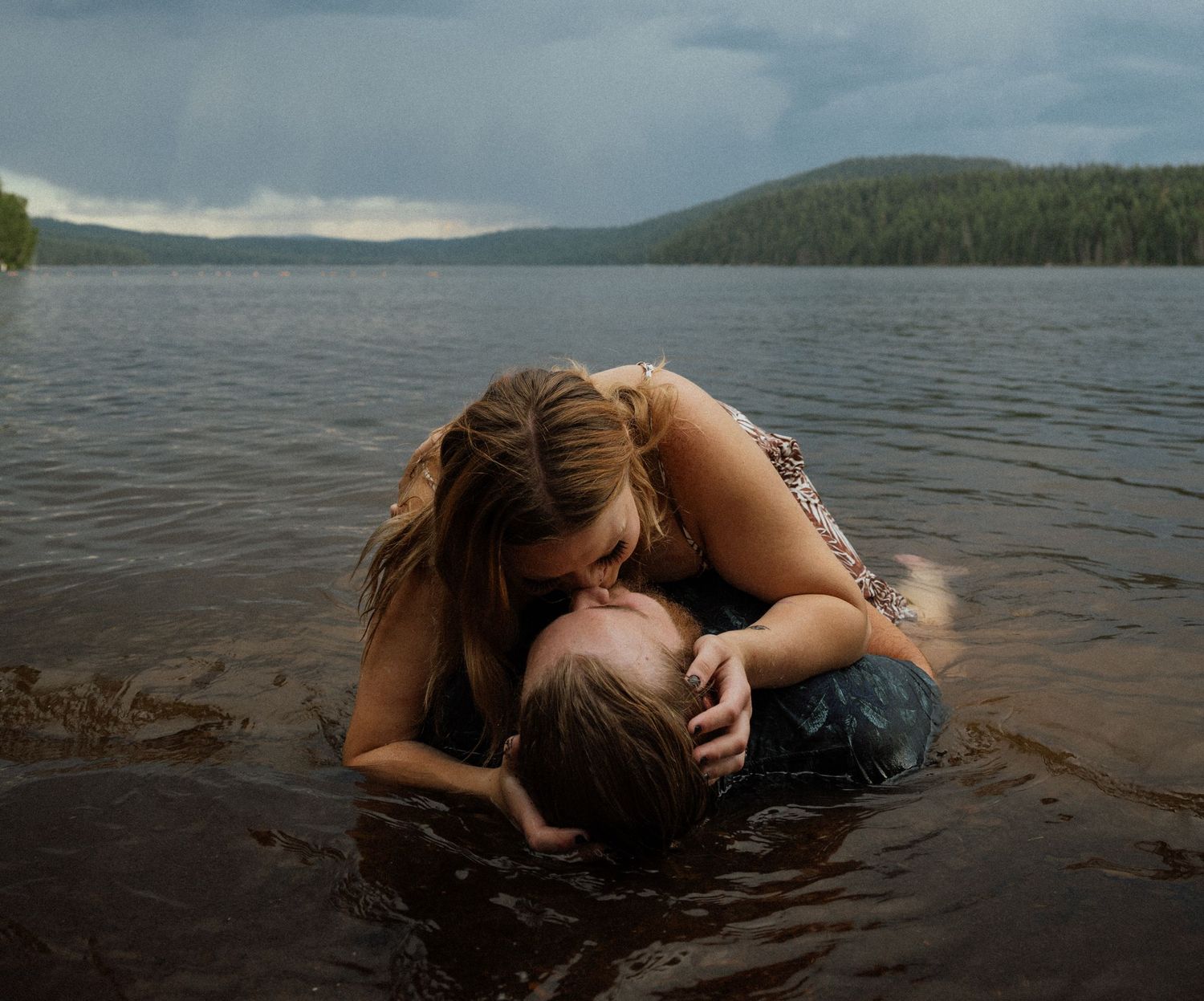 Two people embracing in shallow lake water with stormy skies and mountains in the background.