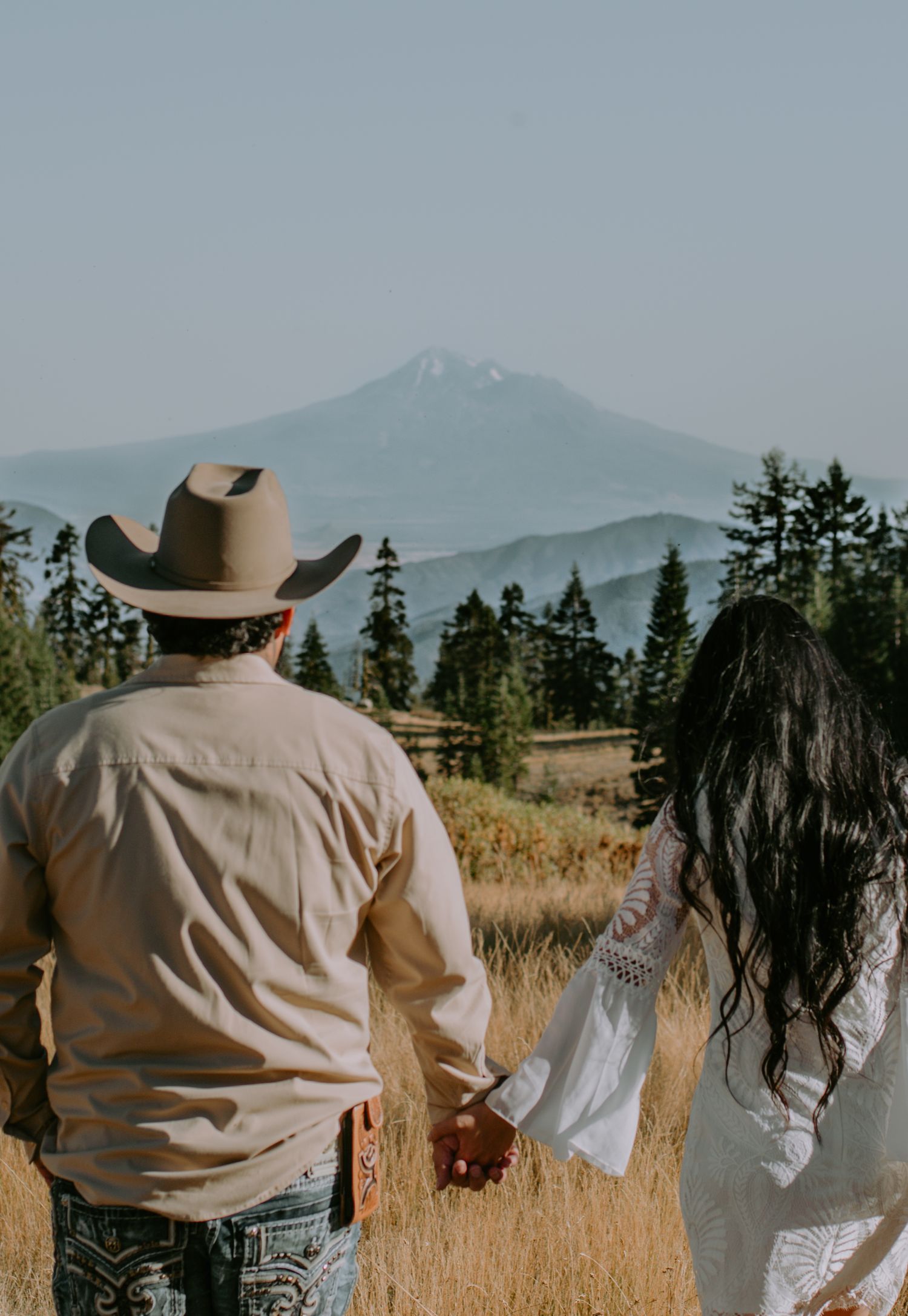 Three people walking through a sunlit meadow with mountains and pine trees in the distance.