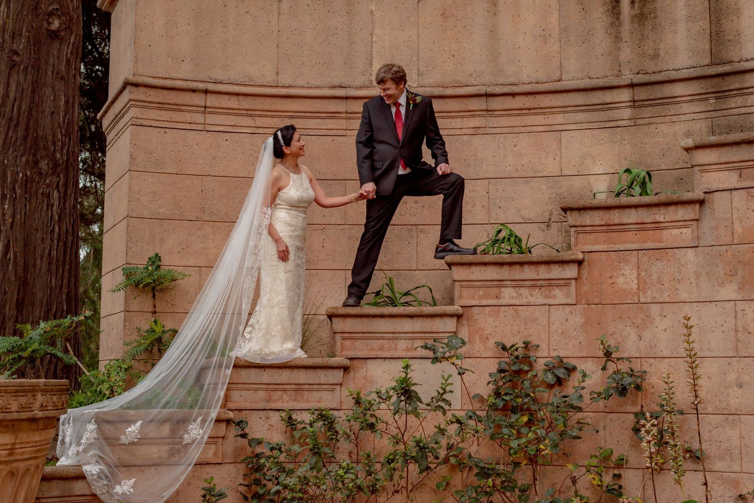 Wedding couple poses along stone architectural wall with trailing veil in romantic portraits.