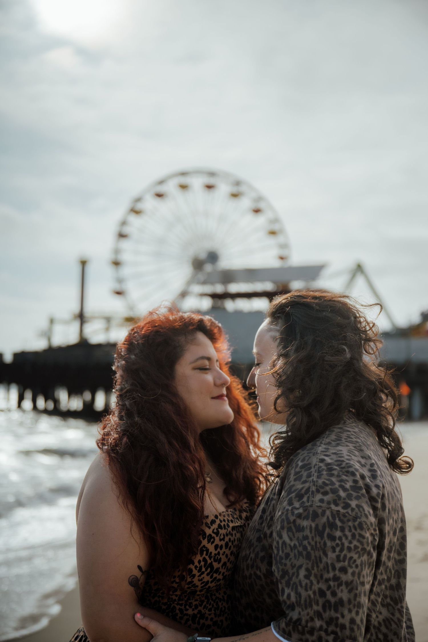 A romantic moment captured at a beach pier with a ferris wheel silhouetted against cloudy skies.